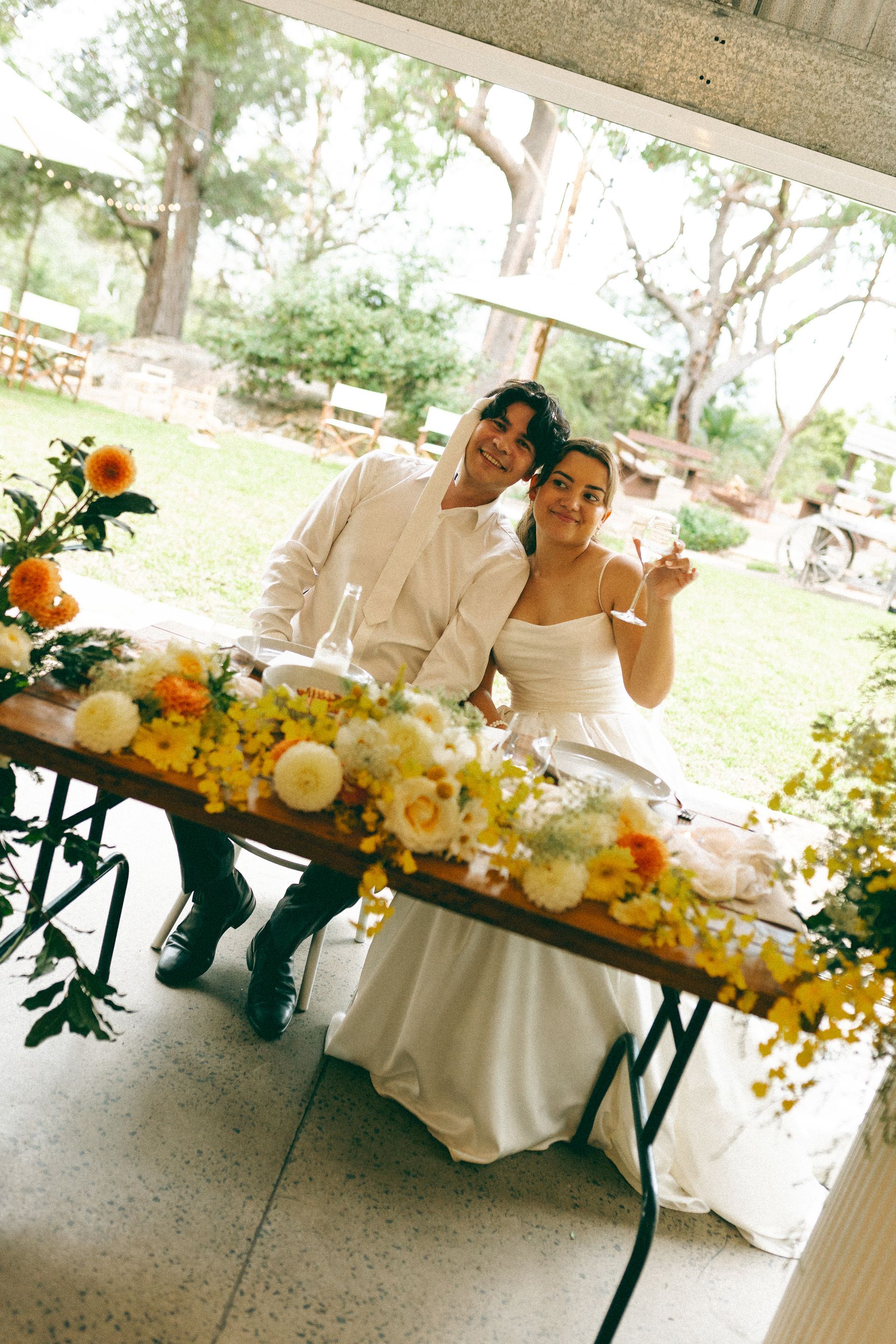 Two people seated at a flower-decorated wedding table under a tent, smiling and posing outdoors.