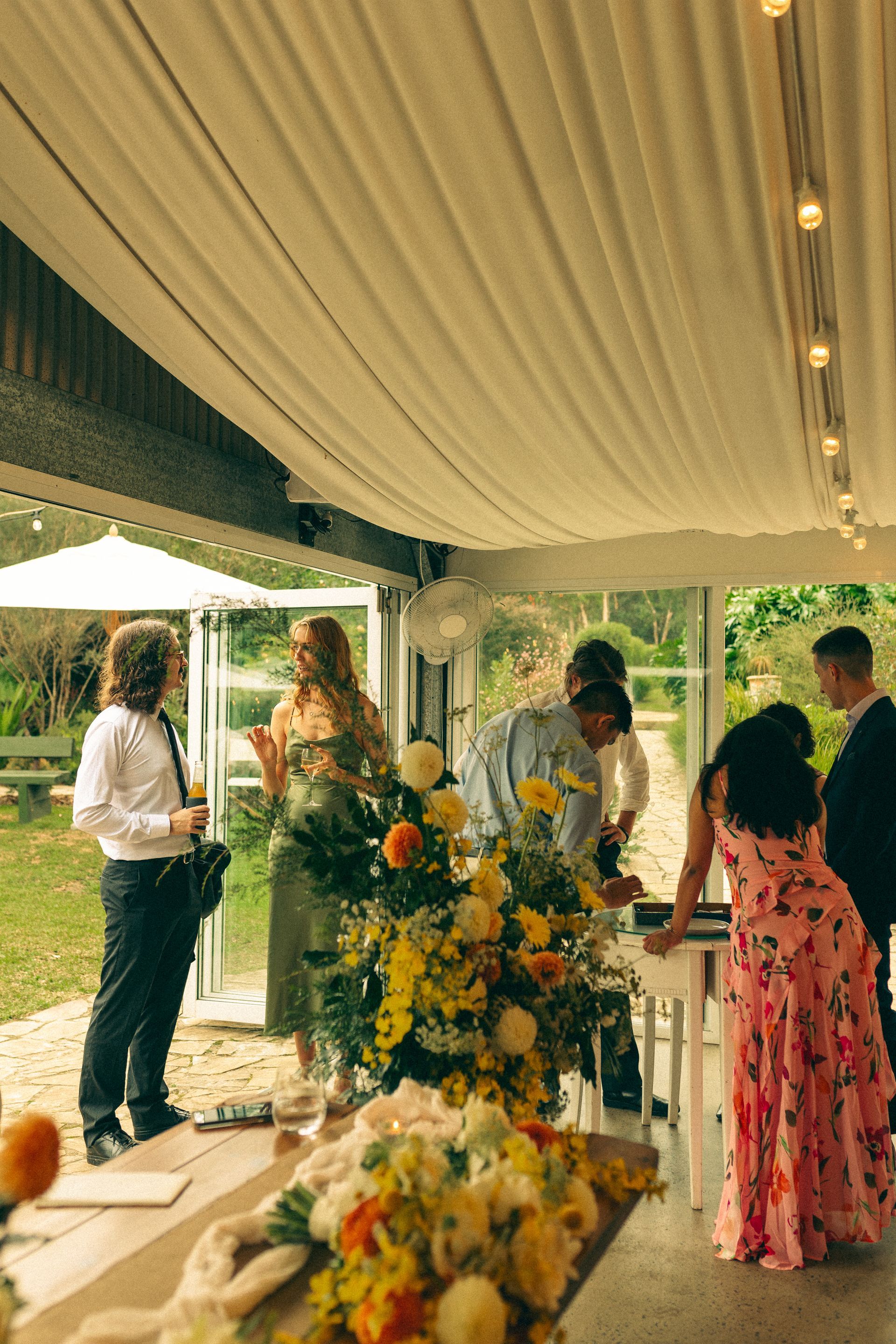 Guests mingle at a floral-decorated outdoor reception under a draped canopy.