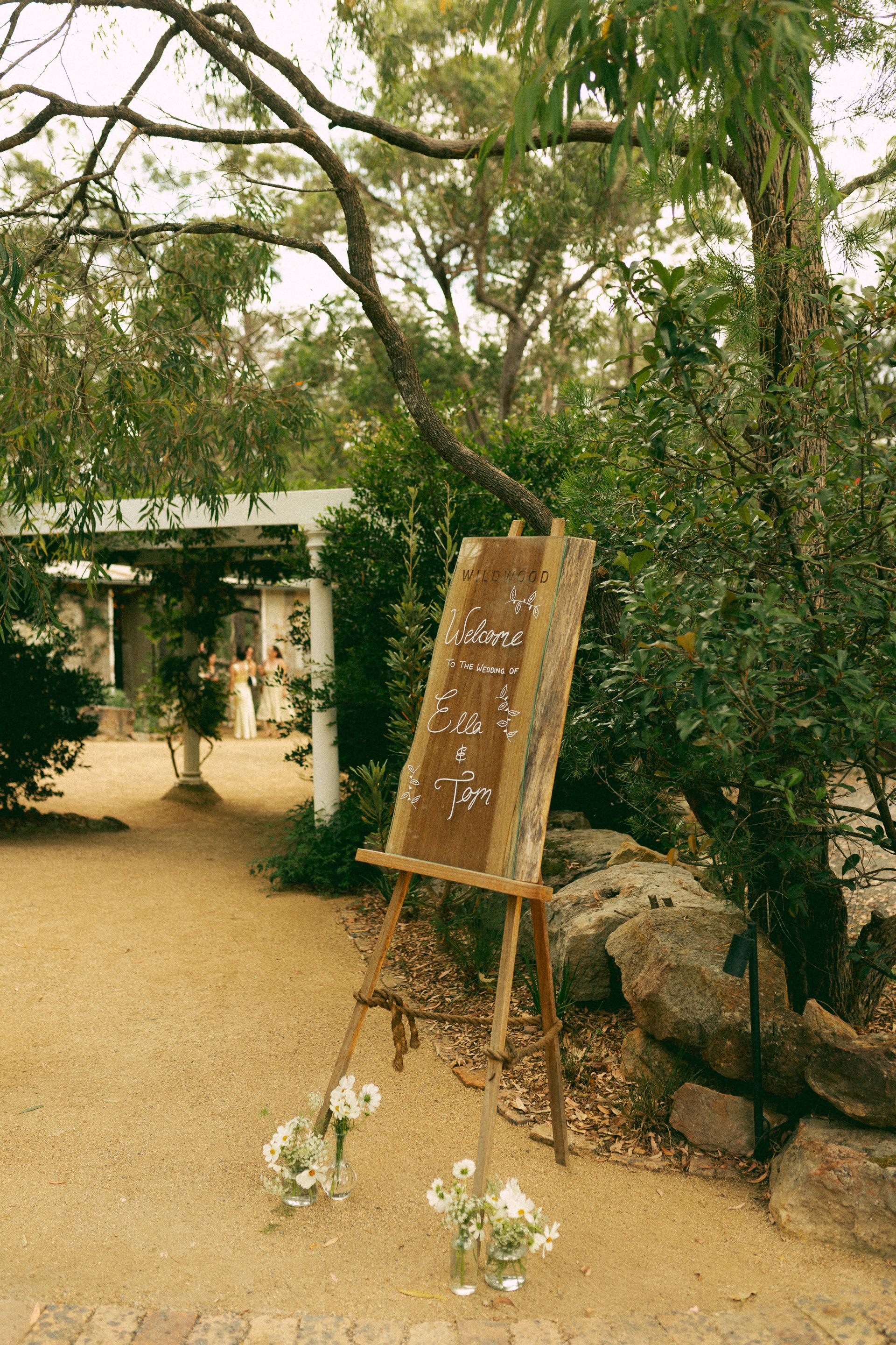 A wooded memorial site with flowers, a signboard, and a small white building in the background