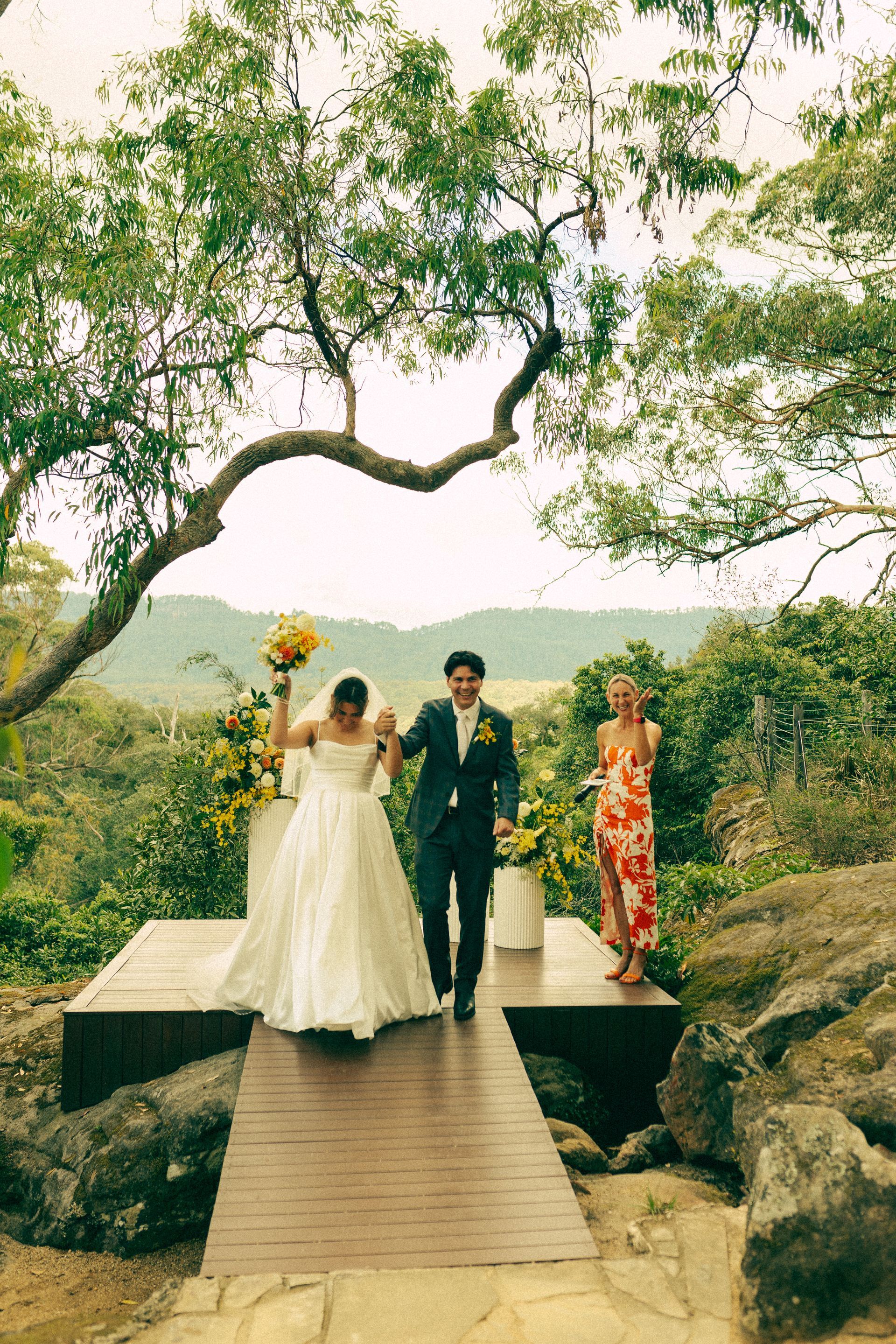 Bride and groom walk down a wooden aisle after their outdoor wedding. A woman looks on. Greenery surrounds them.