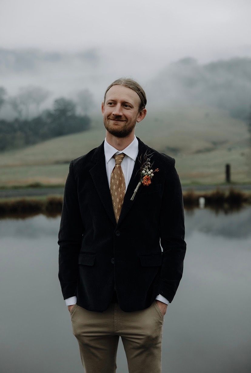 Man in black blazer and patterned tie stands near water, looking to his left; foggy landscape in the background.