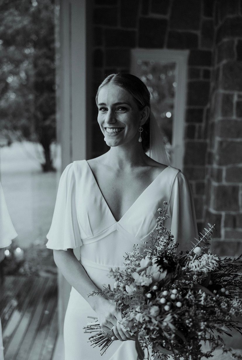 Bride in white dress smiles, holding flowers, standing in a doorway. Black and white photo.