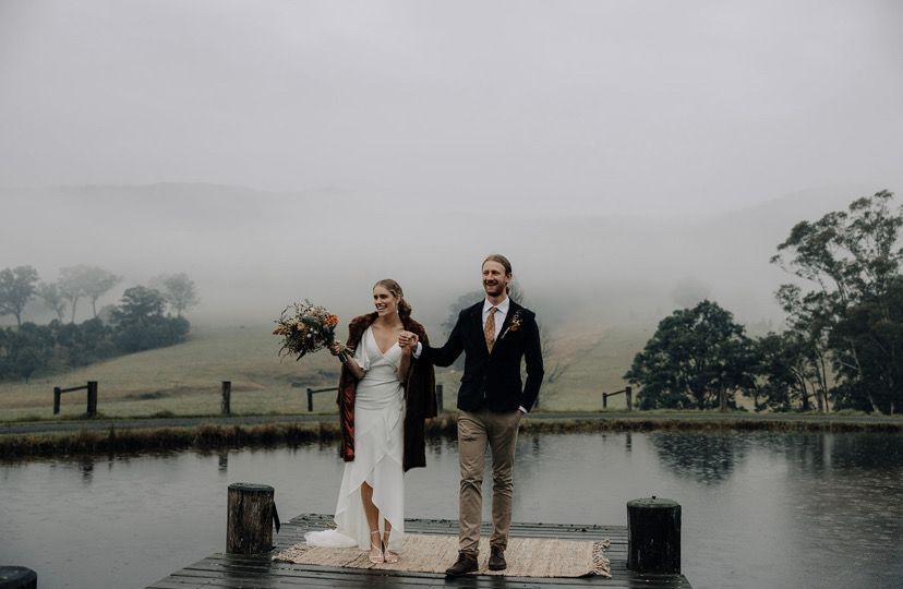 Newlyweds on a dock in the rain. Bride smiles, holding flowers, groom in suit. Foggy lake and hills in background.