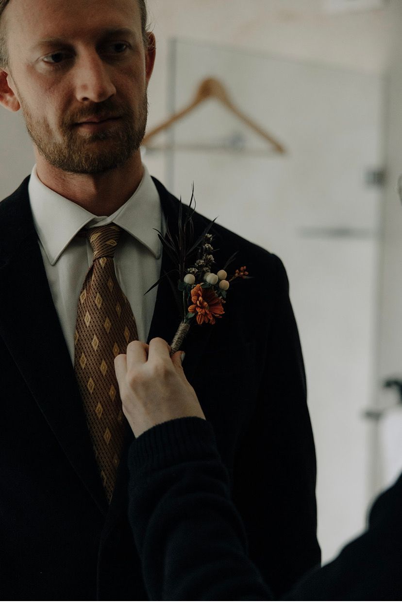 Man in suit has boutonniere adjusted by someone. Reflective mirror, neutral colors.