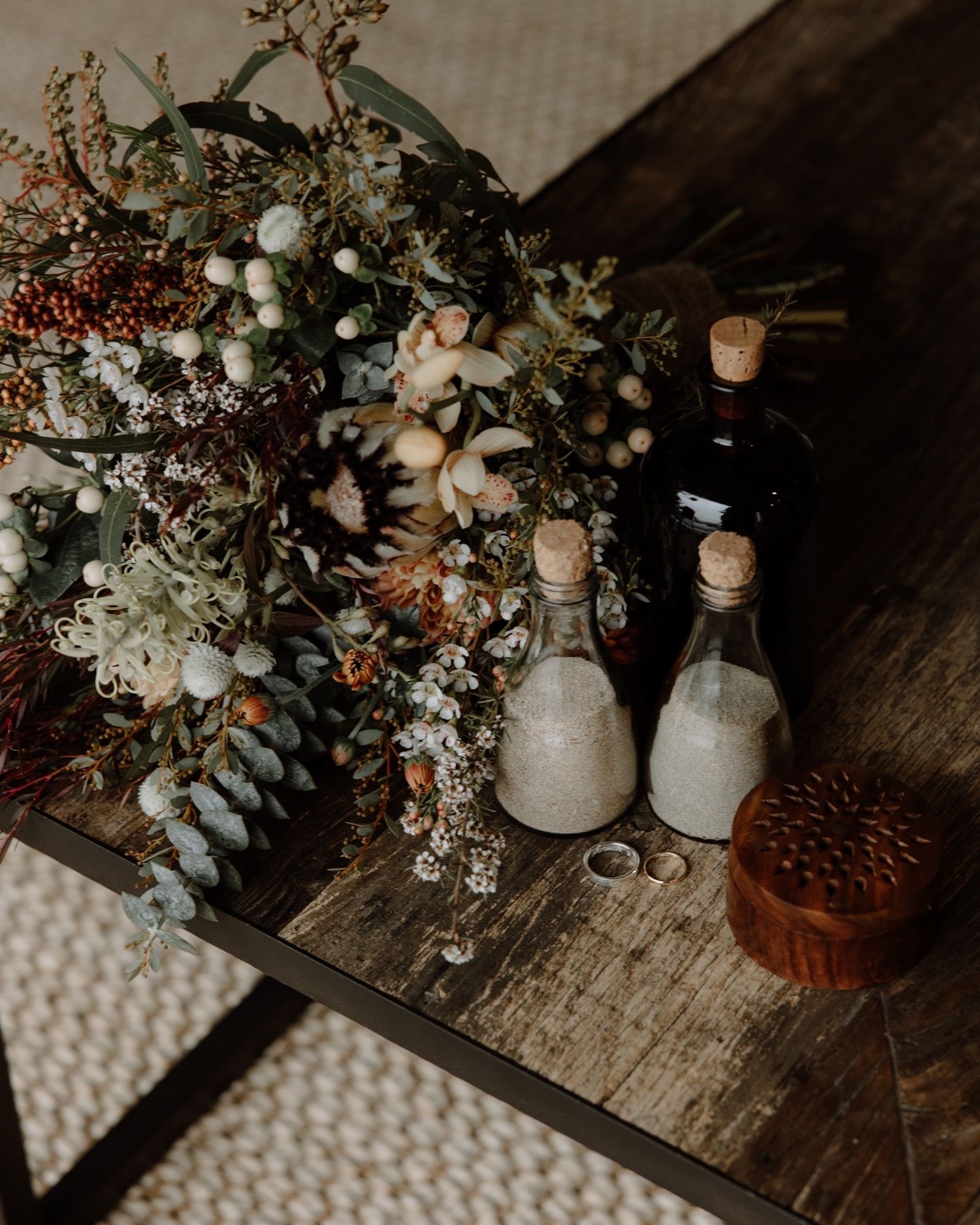 Wedding bouquet, sand ceremony bottles, rings, and wooden box on a rustic wooden table.