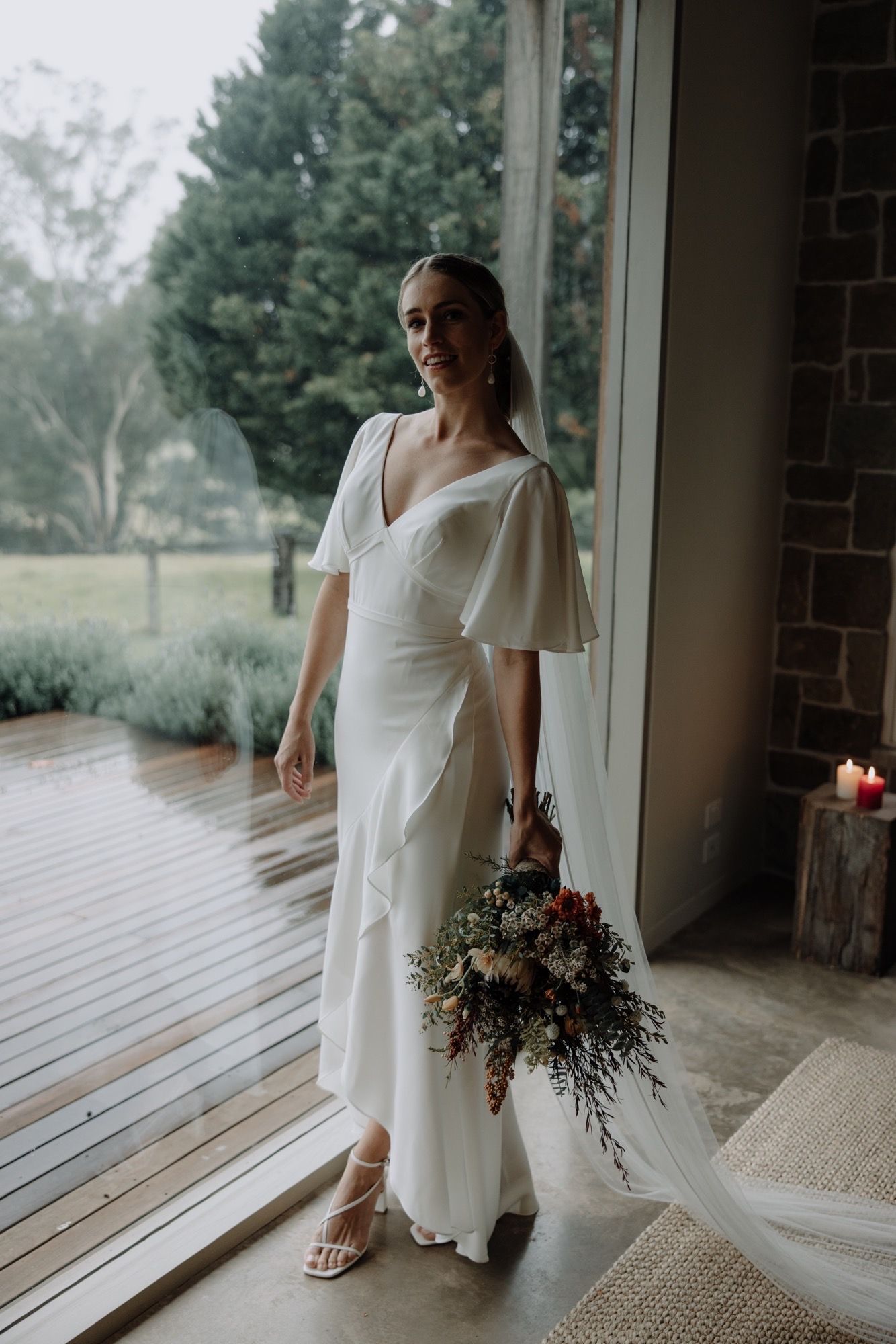 Bride in a white dress, holding flowers, standing by a window with a rainy view.