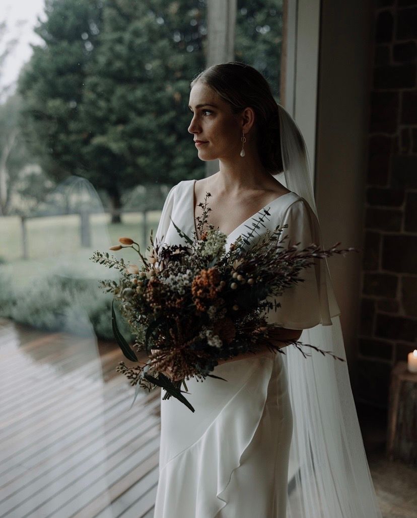 Bride in a white dress holding a bouquet, looking out a window. Rainy day, natural light.