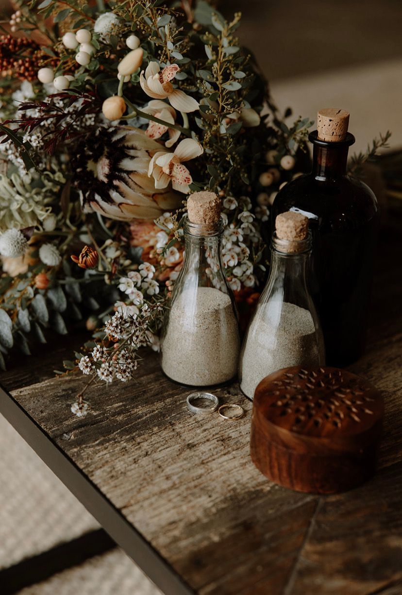Wedding details: bouquet, sand ceremony bottles, rings, wooden box on rustic table.
