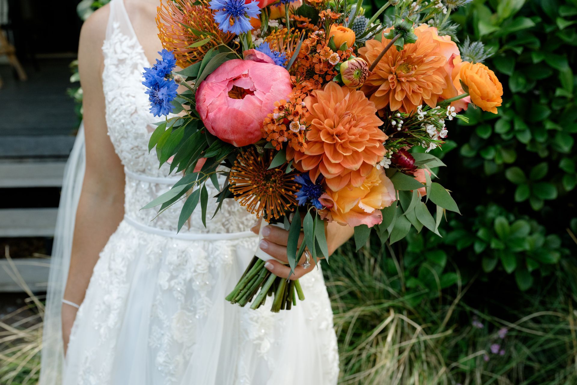 Bride in white lace dress holding a colorful bouquet of orange, pink, and blue flowers outdoors