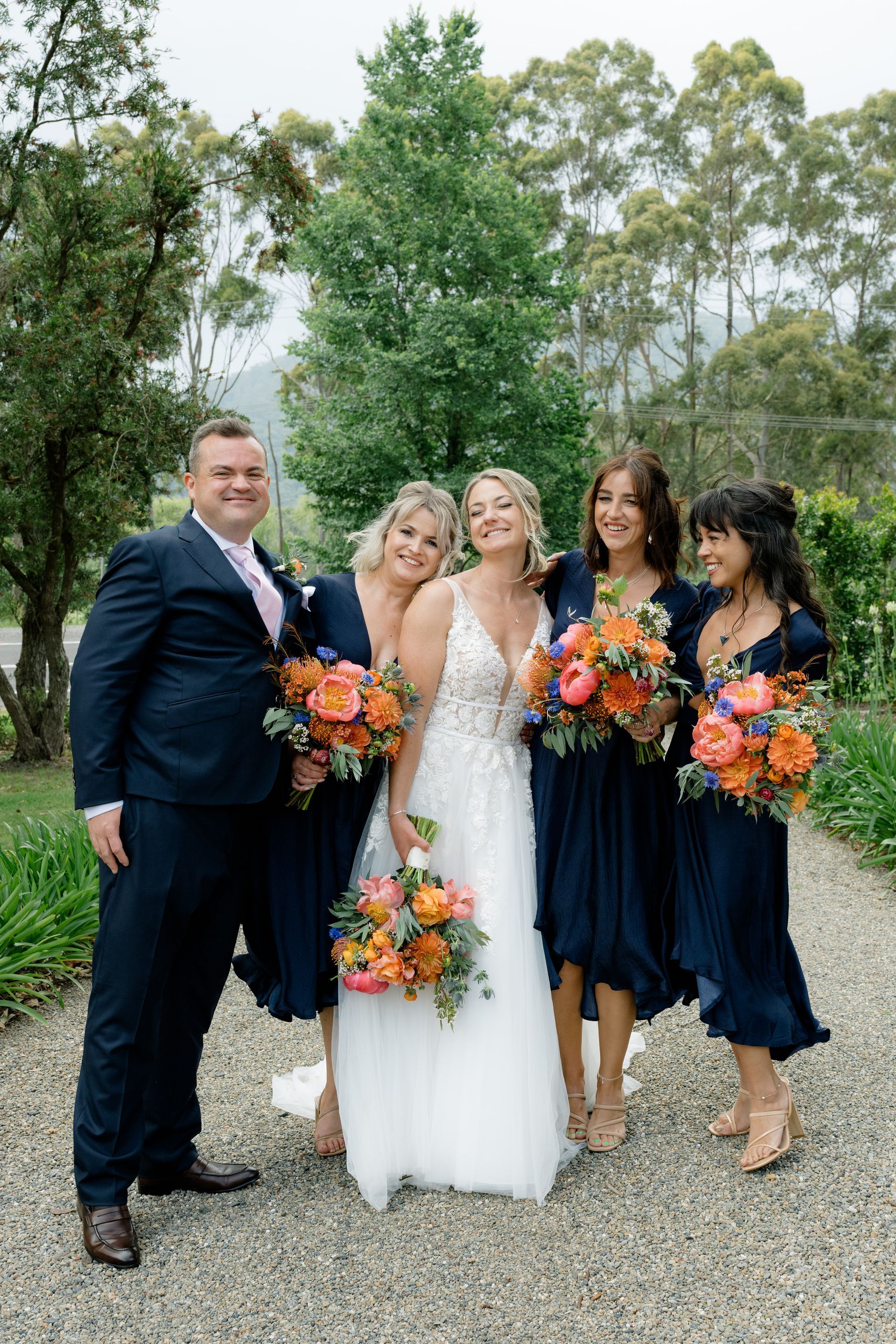 Wedding party posing outdoors, bride in white dress with bridesmaids holding orange and blue bouquets.