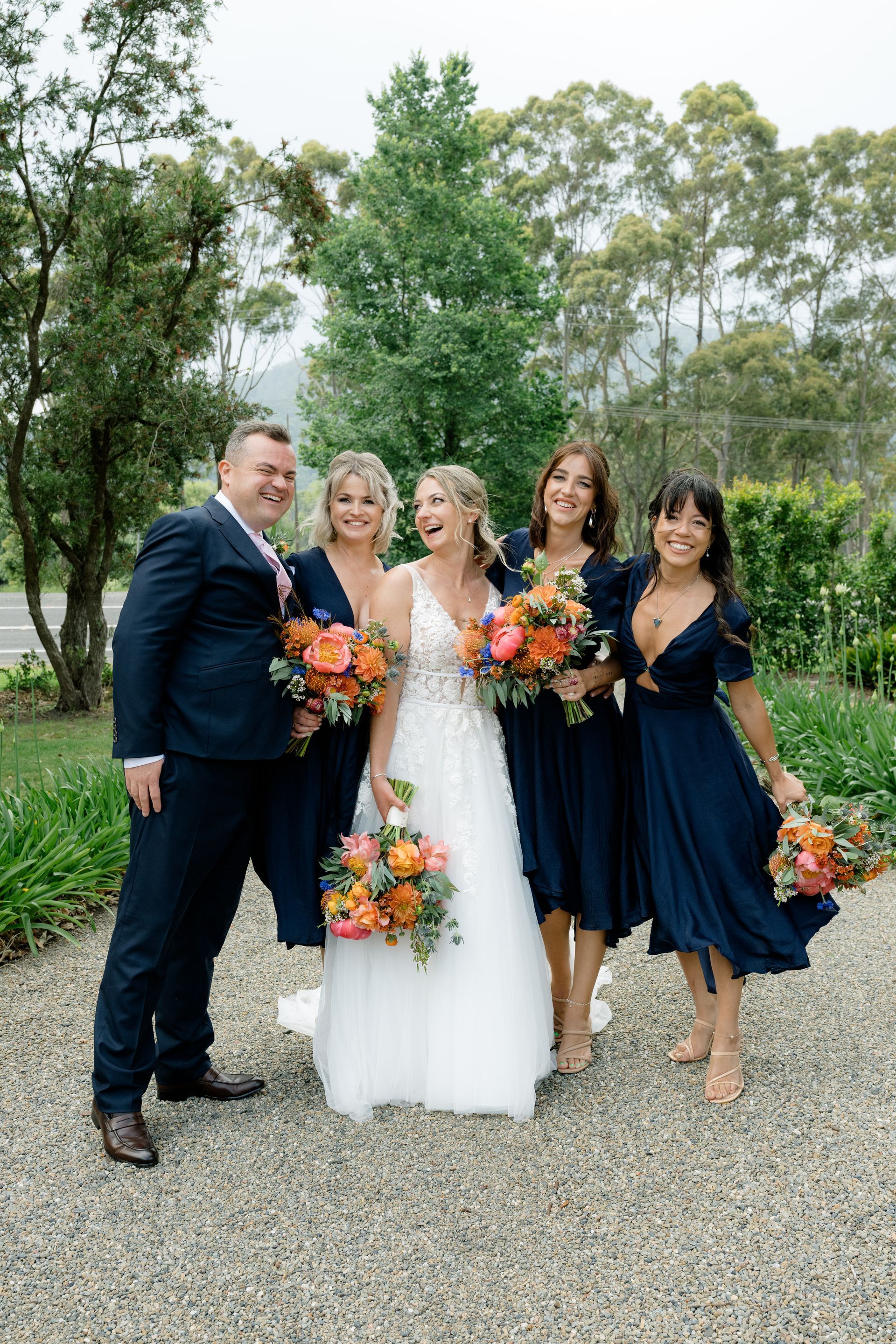 Five people in formal attire posing outdoors, holding orange and pink bouquets on a gravel path.