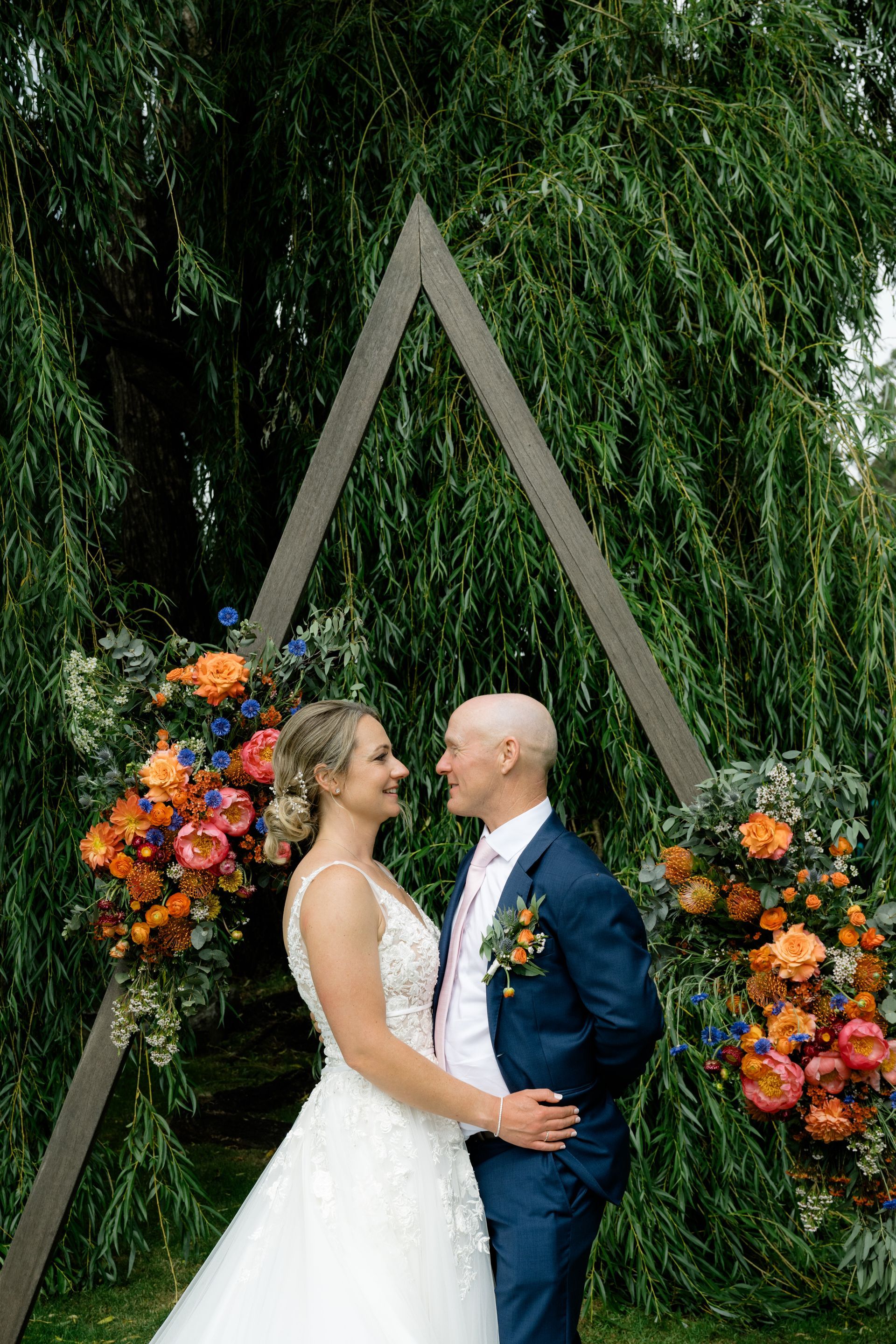 Bride and groom kissing under a triangular floral arch with greenery backdrop