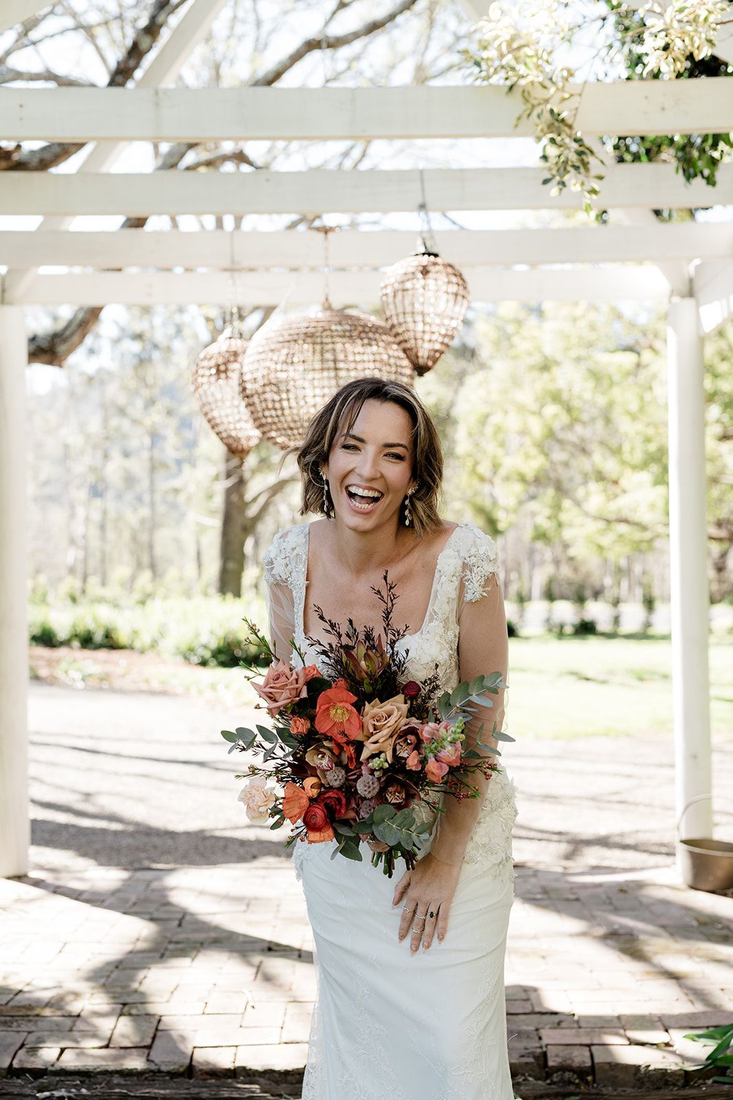 Smiling bride holding a bouquet under a white pergola in a sunny garden.