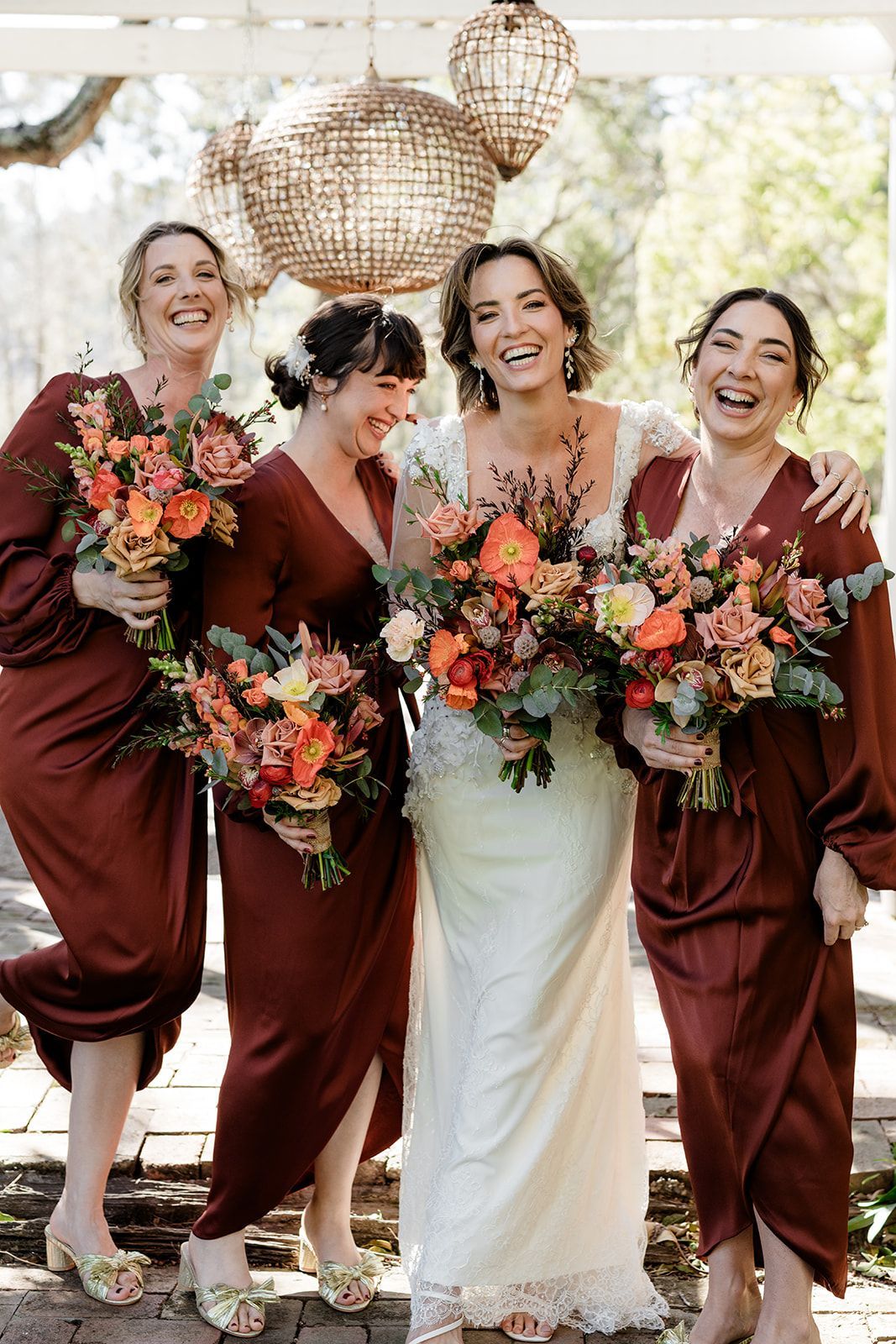 Four women in burgundy outfits and a white dress hold orange bouquets at an outdoor wedding arch