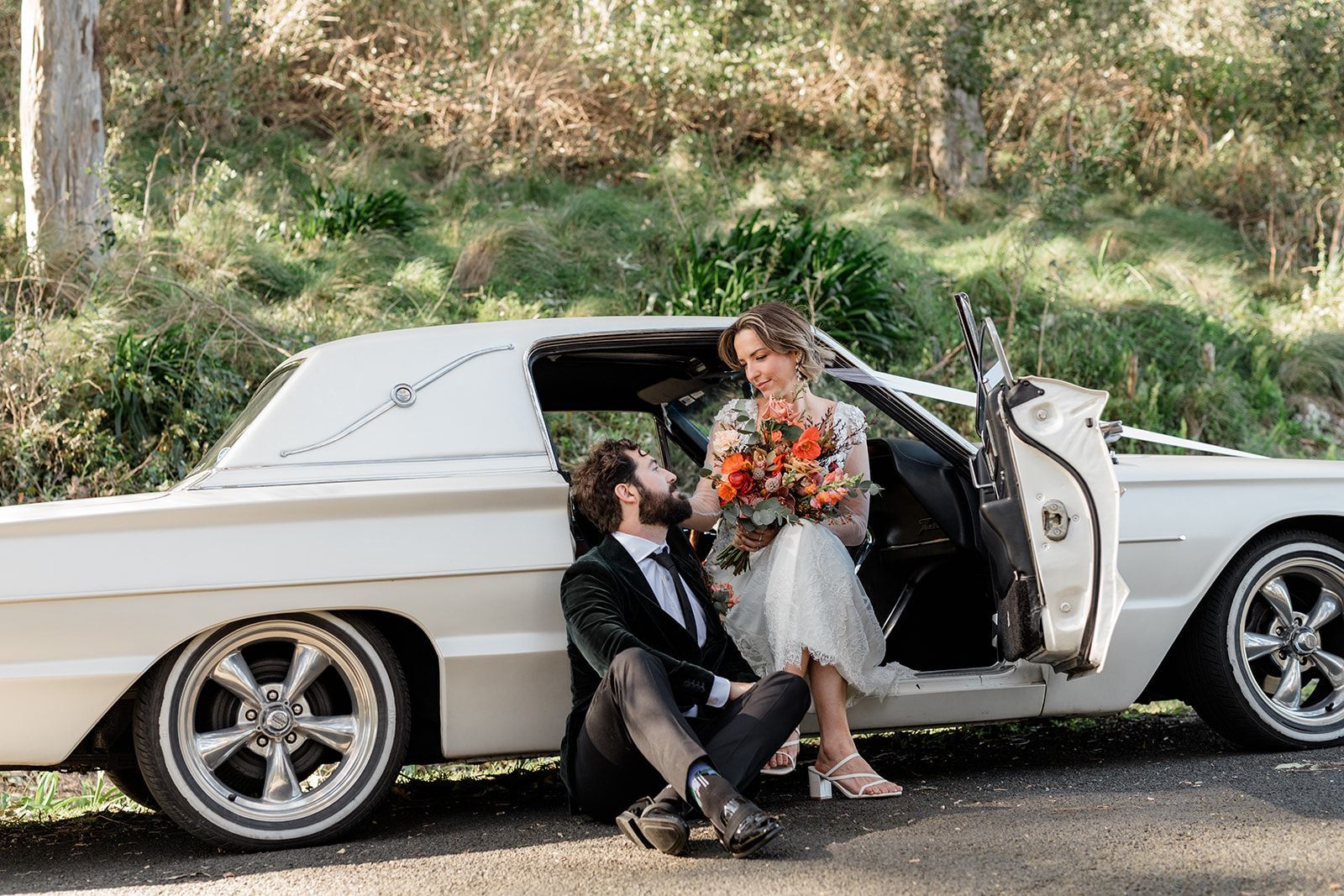Wedding couple by white vintage car, bride stepping out with bouquet as groom kneels beside her.