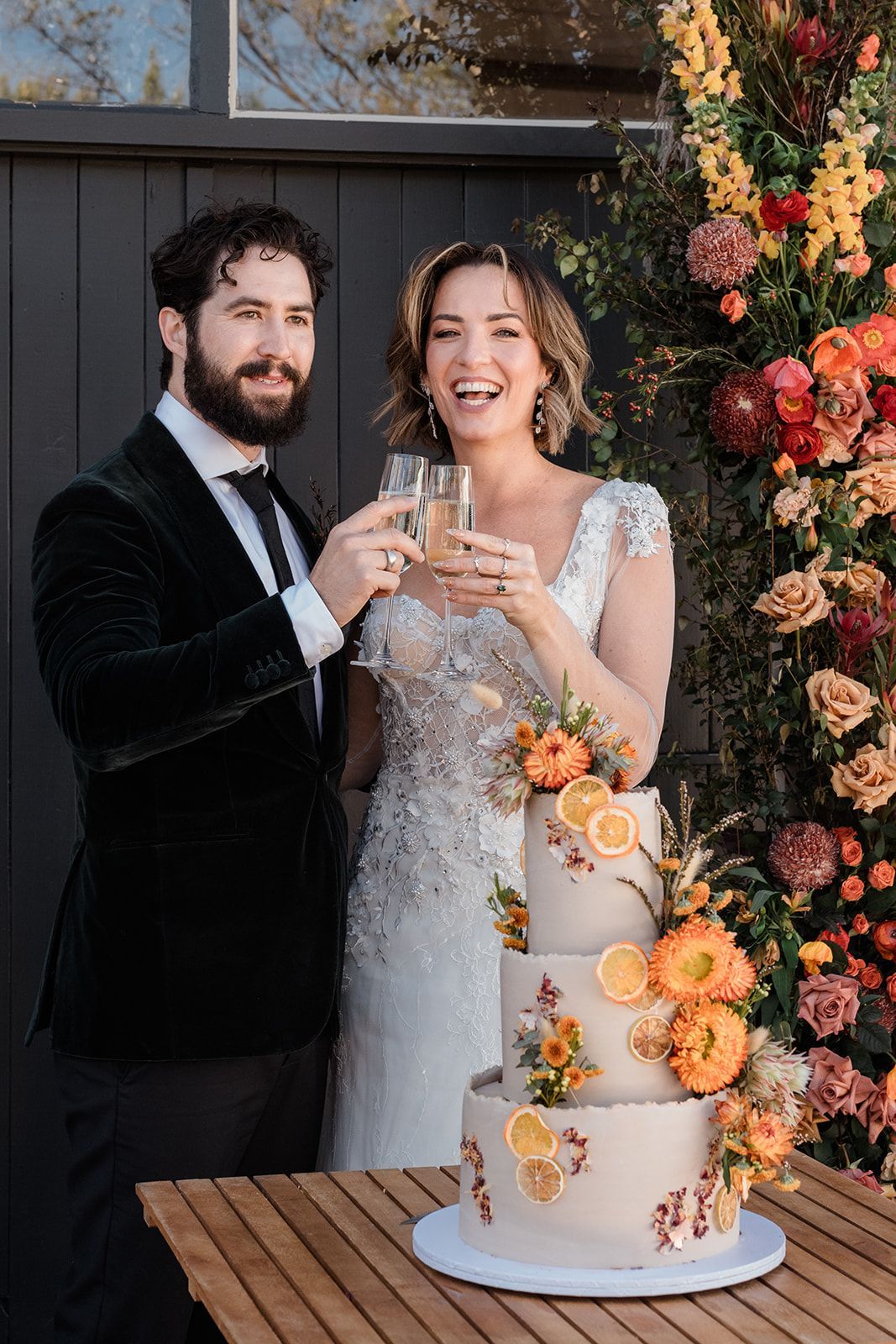 Bride and groom toast beside a three-tier wedding cake decorated with orange flowers.