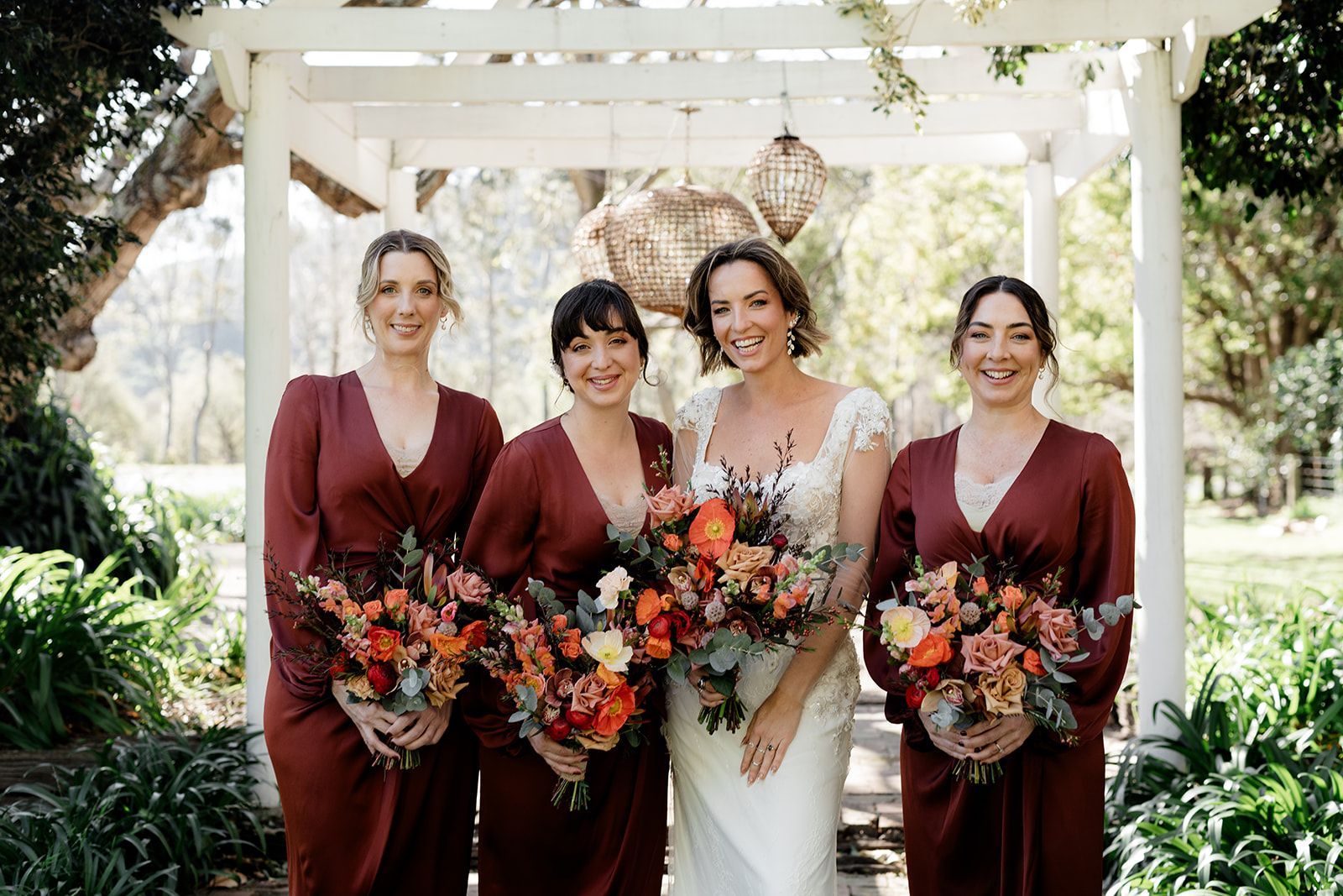 Four women in burgundy dresses and a white gown hold autumn bouquets under a garden pergola.