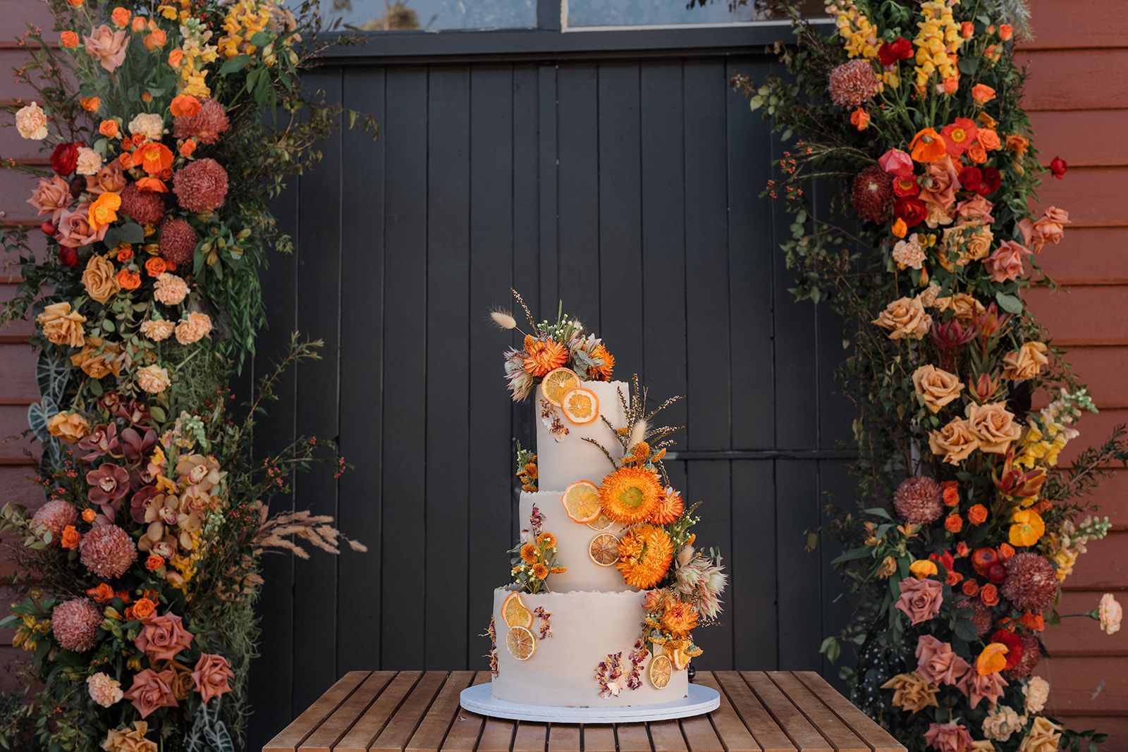 White tiered wedding cake with orange flowers, centered against a dark wall and flanked by floral garlands.
