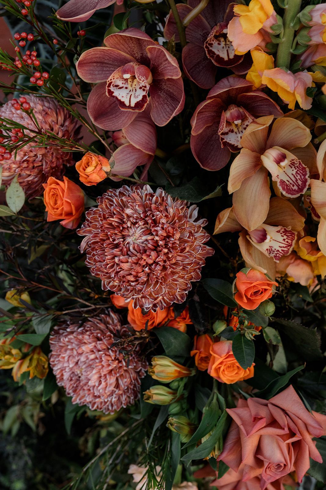 Floral arrangement with burgundy orchids, peach roses, orange blooms, and dark green foliage
