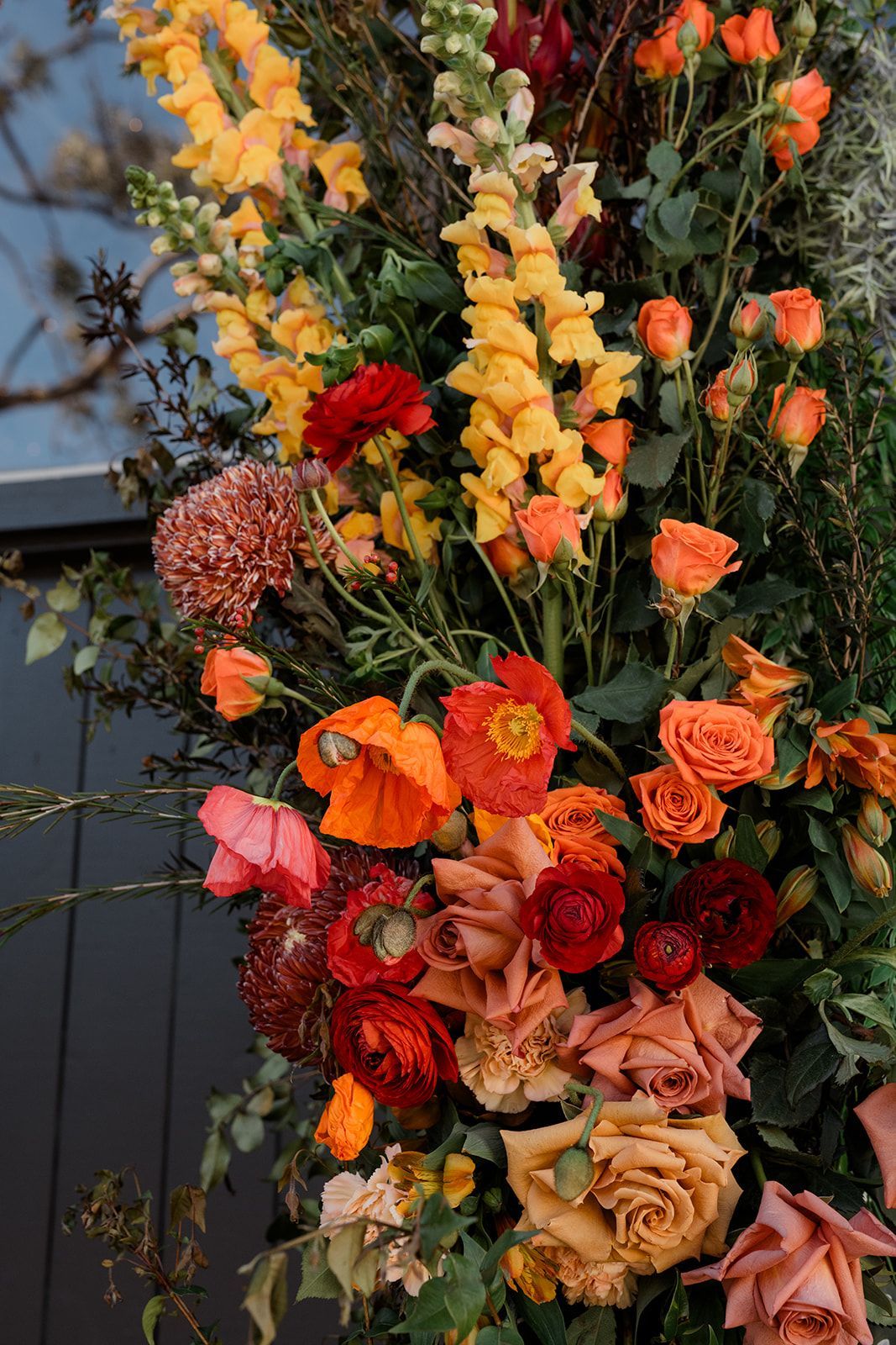 Tall floral arrangement with orange, yellow, pink, and red flowers against an outdoor wall