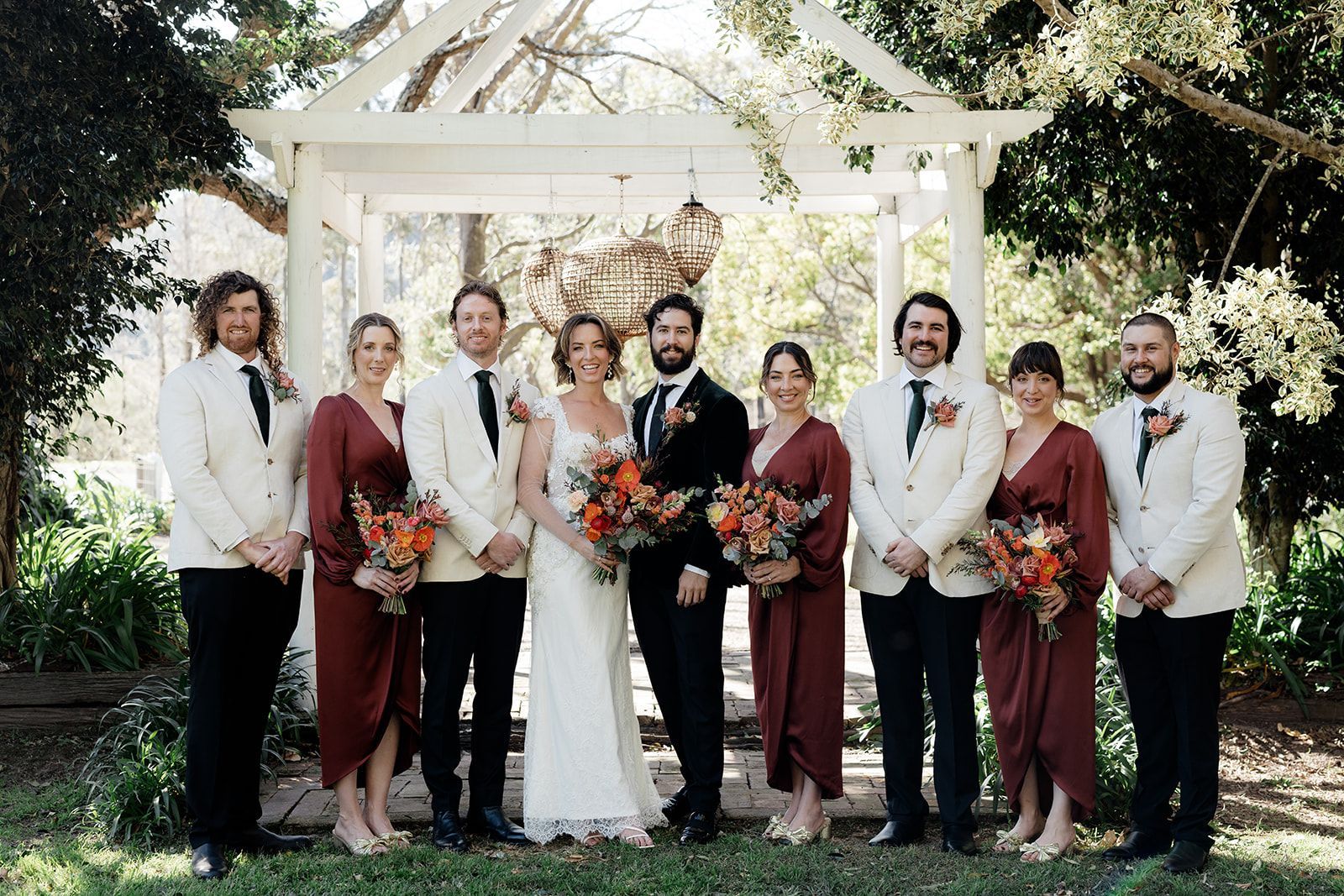 Wedding party posing under a white pergola, holding bouquets in an outdoor garden.