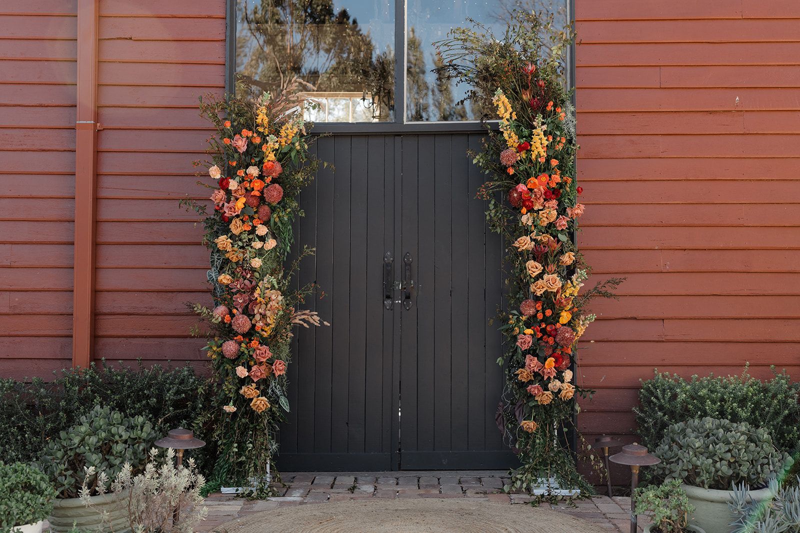 Black double doors framed by tall autumn floral garlands against a red wooden wall.