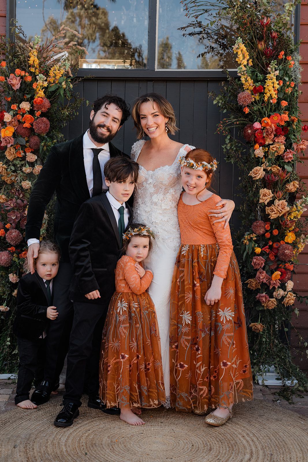 Family of five posing in formal wear before a floral backdrop, with the parents and three children smiling.