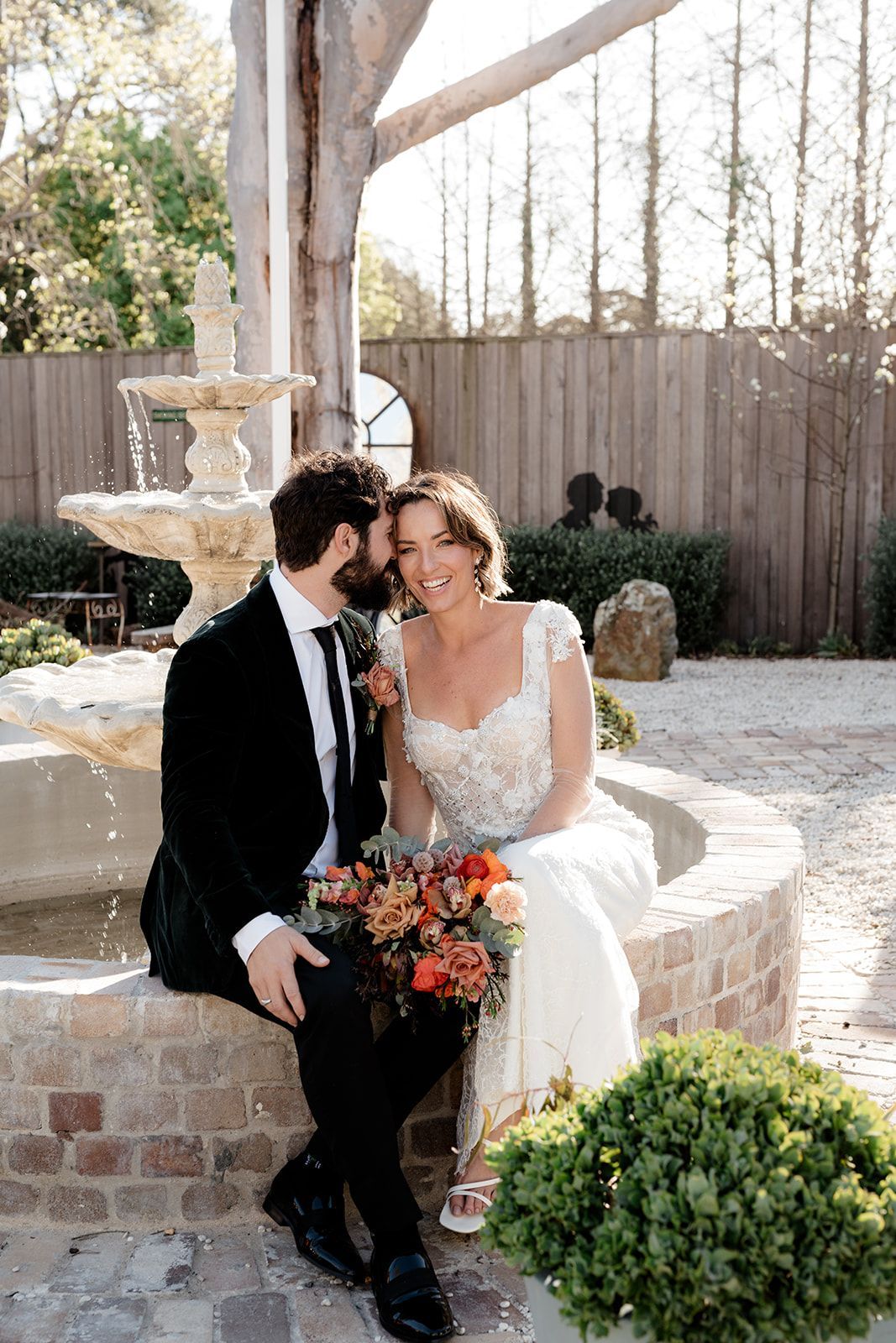 Bride and groom sitting by a fountain, smiling and holding a bouquet outdoors