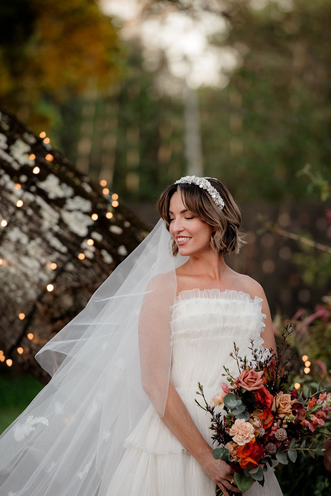 Bride in a white gown holding a colorful bouquet outdoors by twinkling lights.