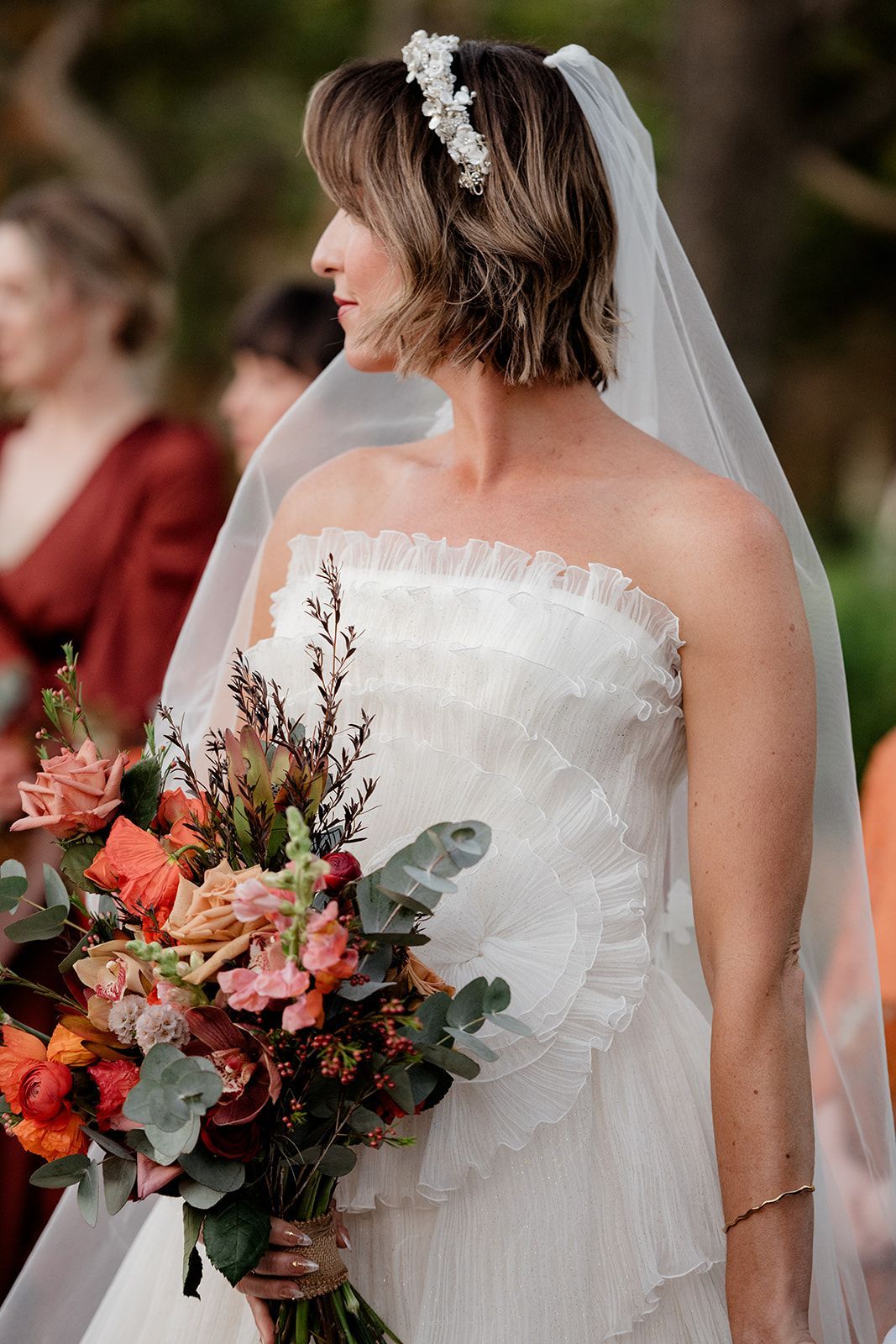 Bride in a strapless white gown holding a colorful bouquet, standing outdoors with a veil and floral headpiece