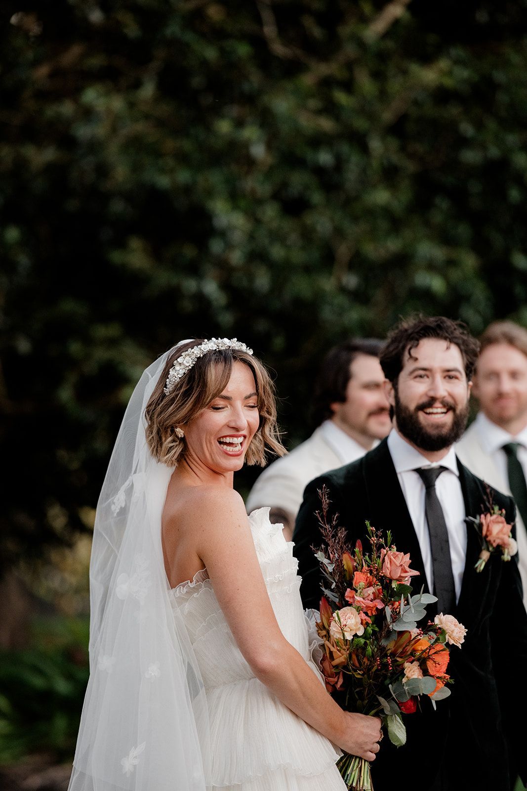 Bride smiling at her groom during an outdoor wedding, holding a bouquet with guests blurred behind them