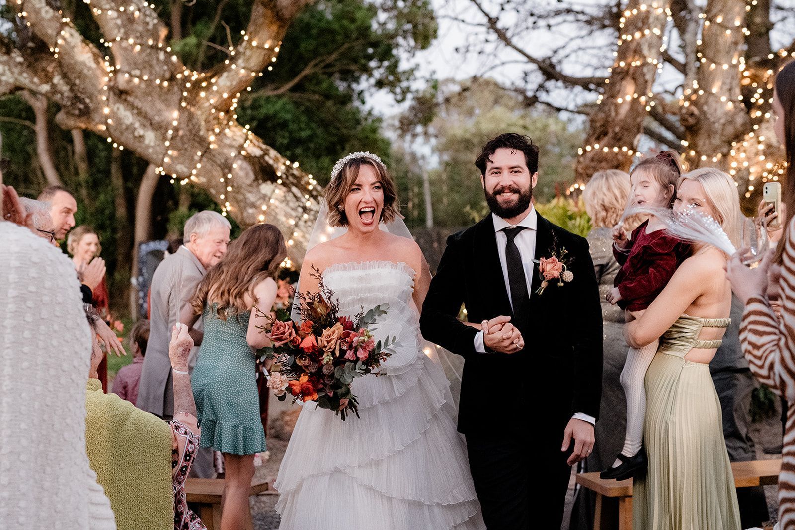 Wedding couple smiling under string lights, bride holding bouquet and groom in black suit with guests nearby