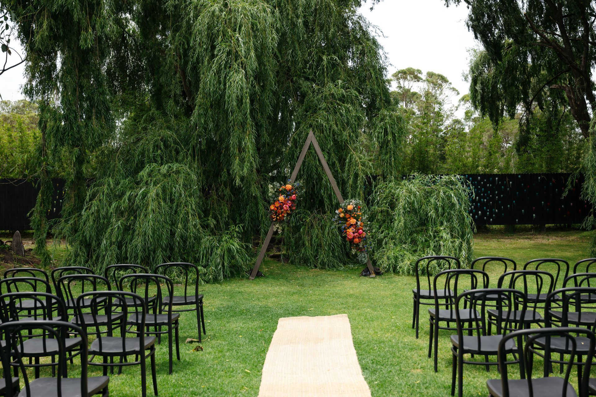 Outdoor wedding aisle with chairs facing a floral arch under a large willow tree
