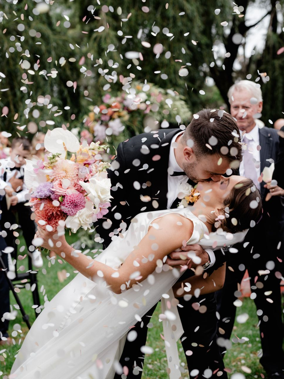 Groom kissing bride, dipped back, amid confetti. Wedding in garden.