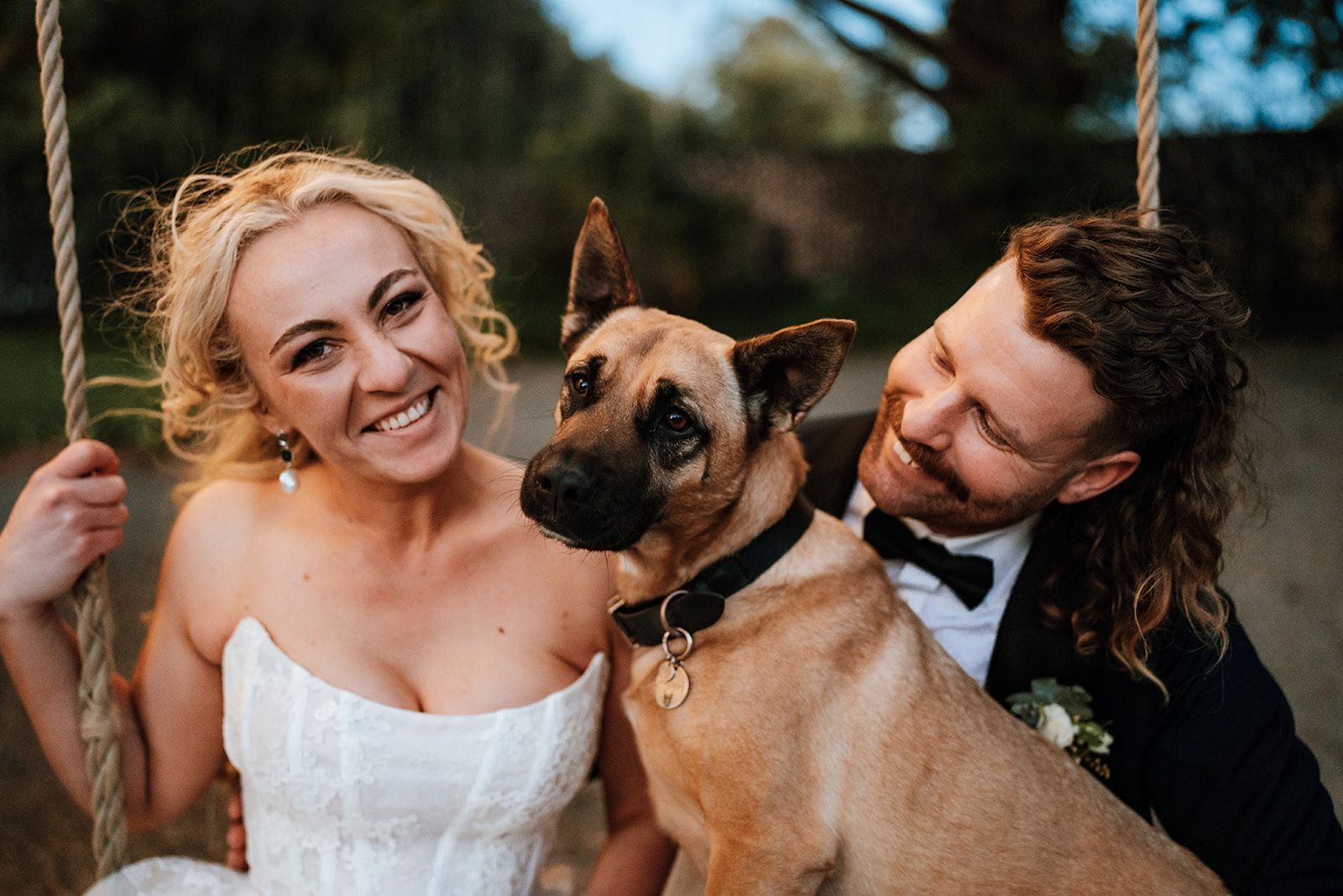 Smiling couple with a tan dog outdoors, the woman in a white dress and the man in a dark suit smiling at the dog