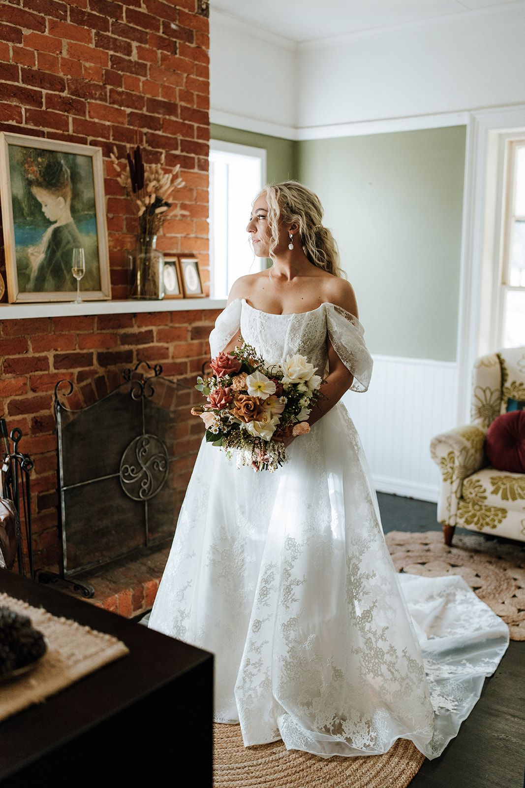 Bride in a white lace gown holding a bouquet beside a brick fireplace in a sunlit room