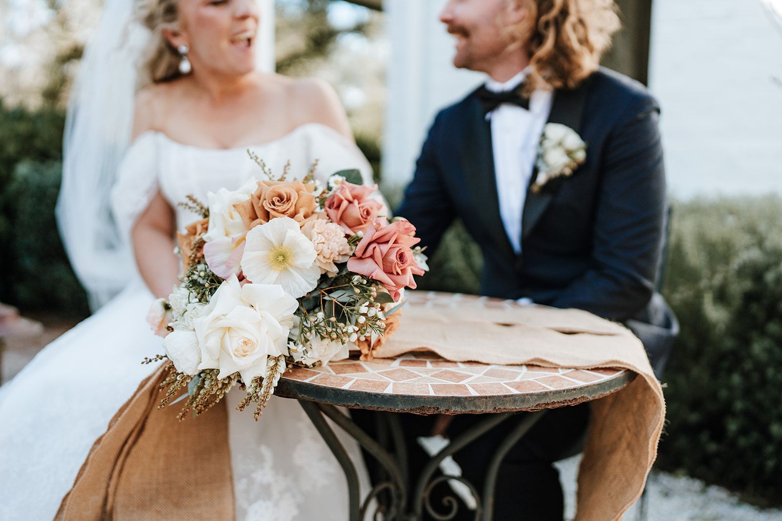 Bride and groom seated beside a rustic table with a pastel floral bouquet outdoors