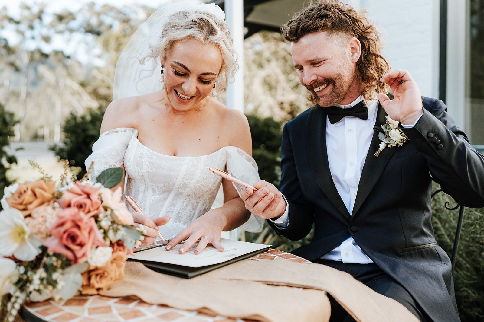 Bride and groom signing a wedding document outdoors beside a bouquet