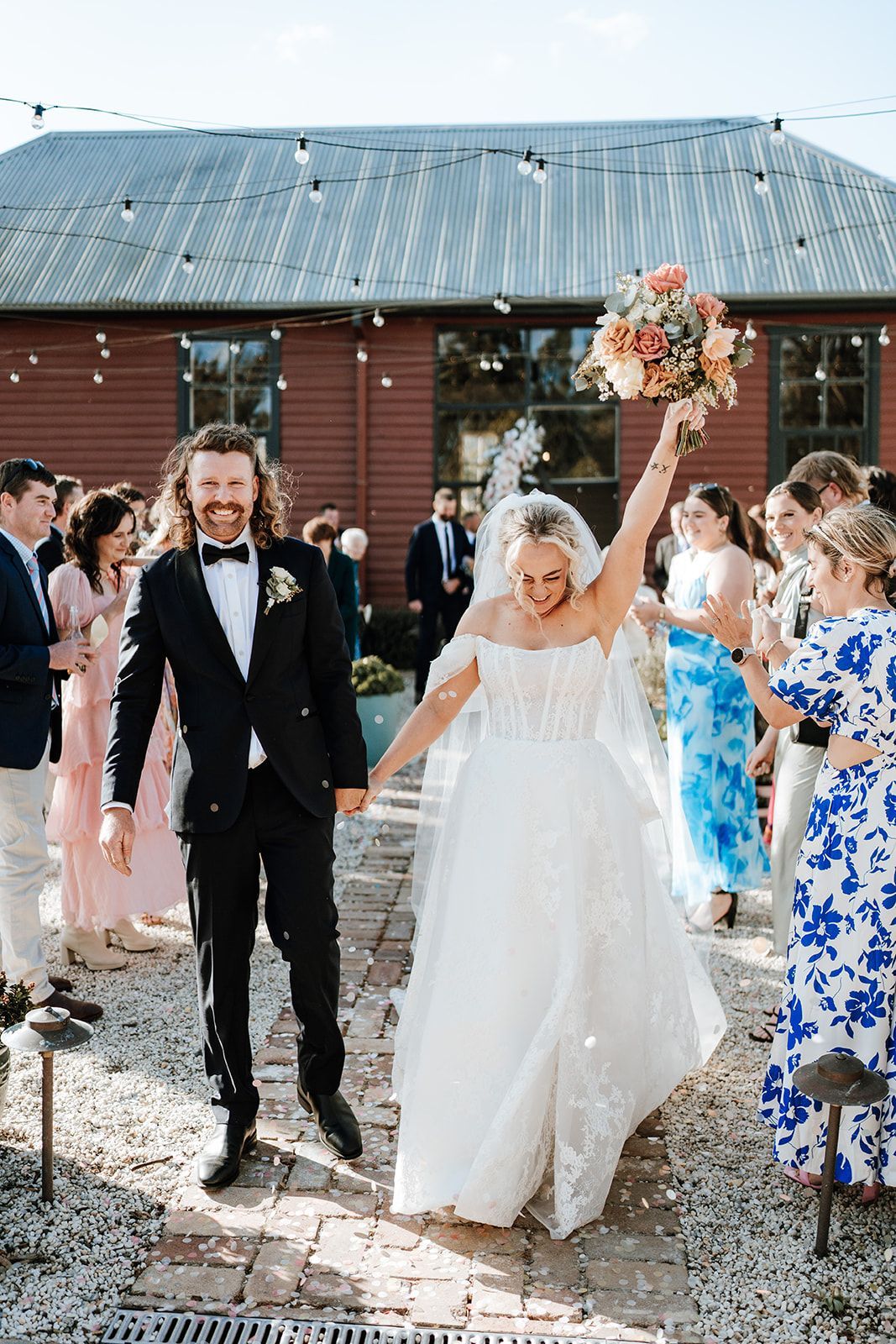 Wedding couple walking down a rustic outdoor aisle, bride raising bouquet, guests cheering near a barn