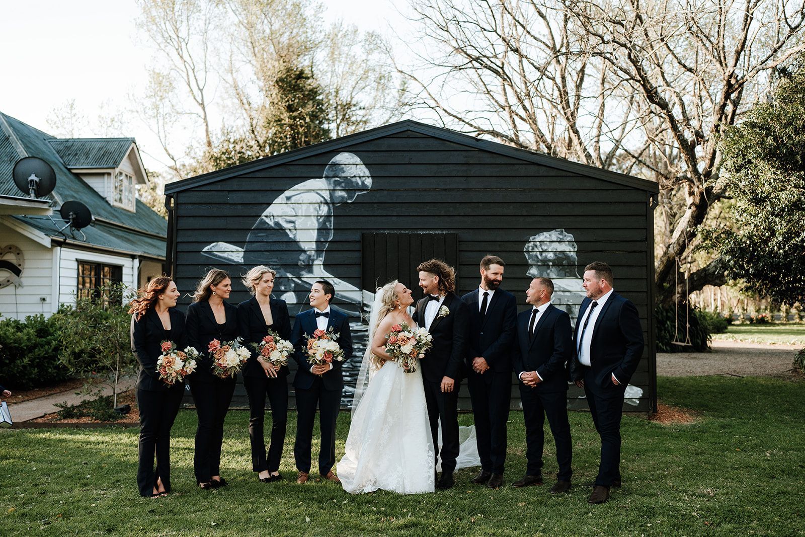 Wedding party posing outdoors in front of a black garage, with the bride in white centered among attendants.