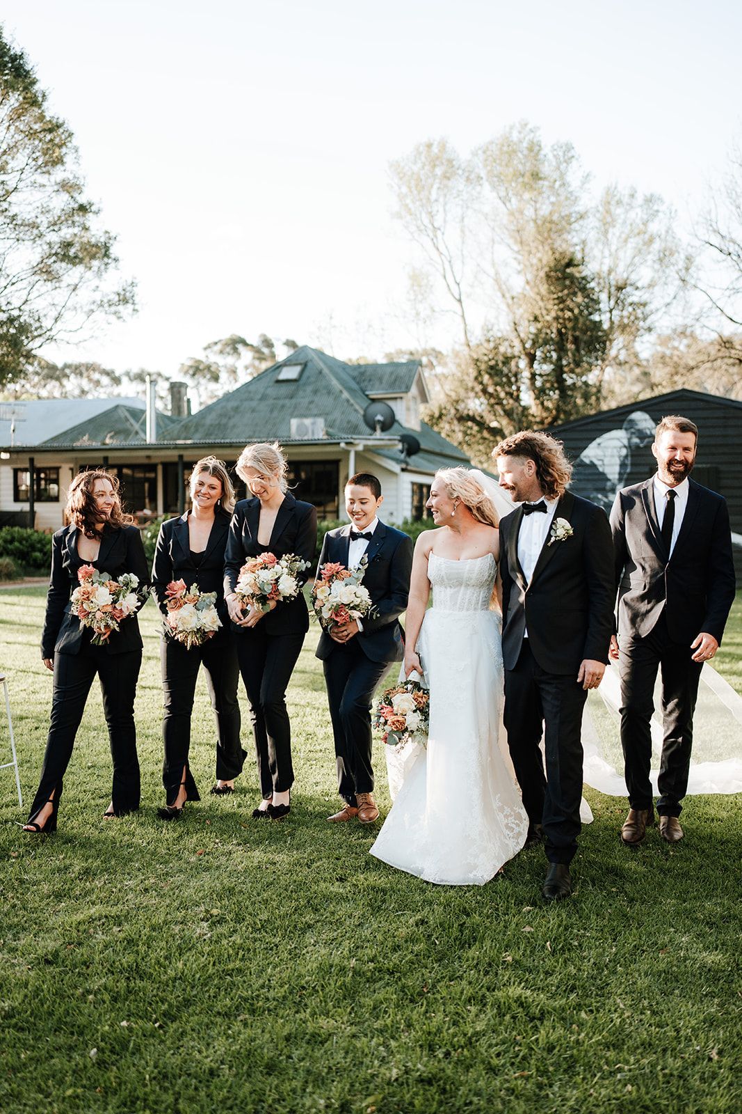 Wedding party posing on a lawn, bride in white dress centered with groom and groomsmen holding bouquets.