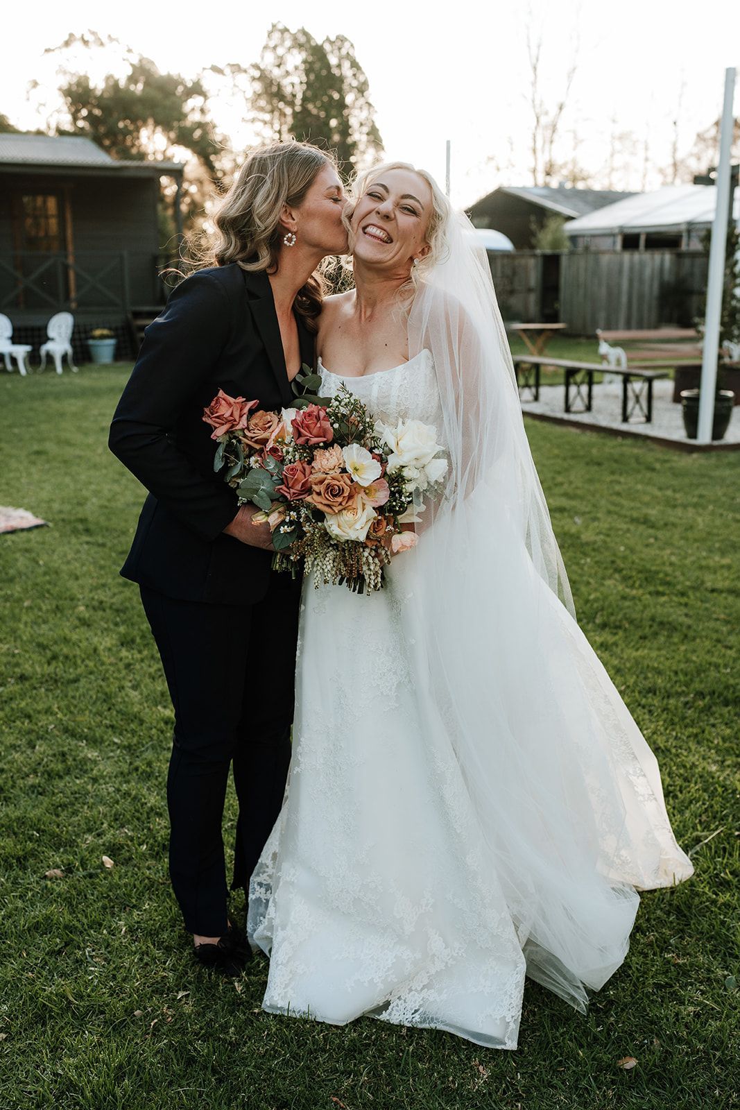 Two brides kiss outdoors, one holding a colorful bouquet in a grassy garden.