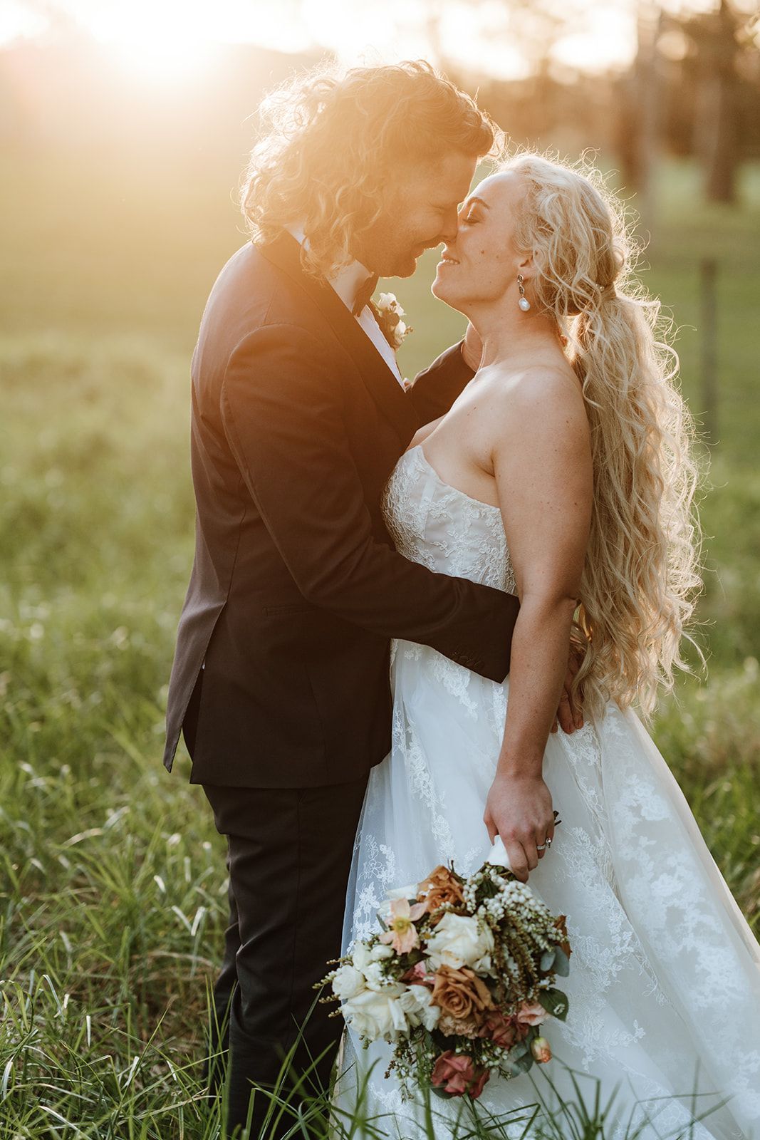 Couple embracing in a sunlit field, bride in white gown holding bouquet, groom in black suit.