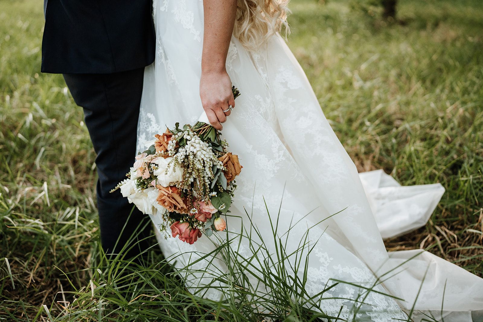 Bride in a white gown holding a bouquet beside a groom in a dark suit on a grassy path.