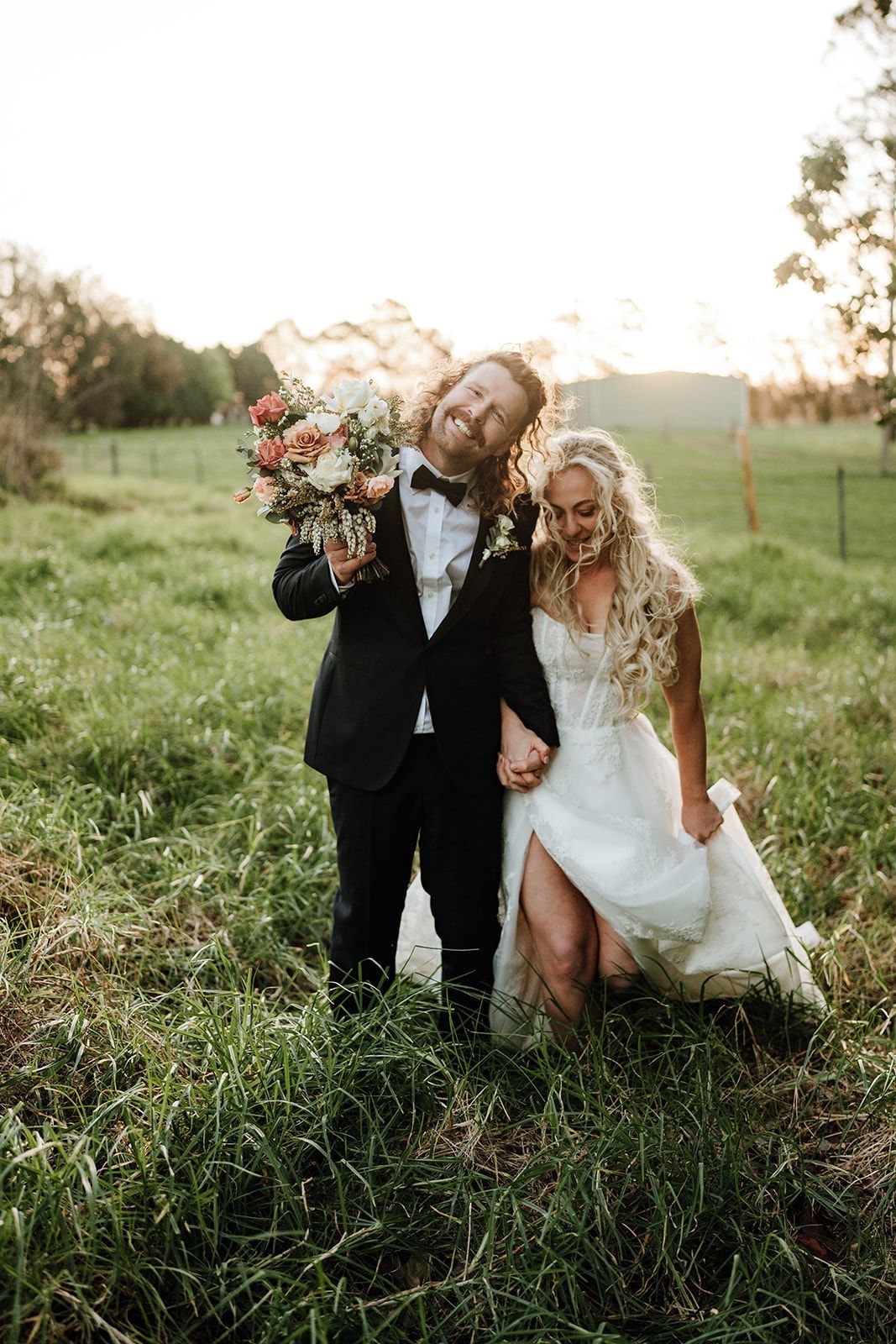 Two people in formal black-and-white attire pose in a grassy field at sunset, holding flowers.