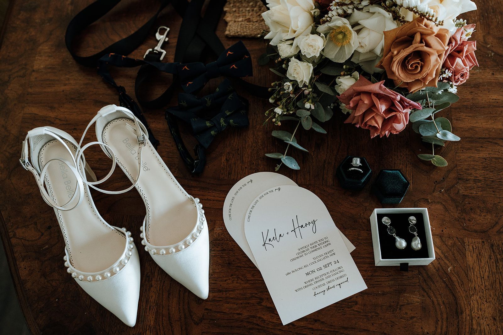 Wedding flat lay with white heels, invitation, rings, black ribbon, and pink-white flowers on a wooden table