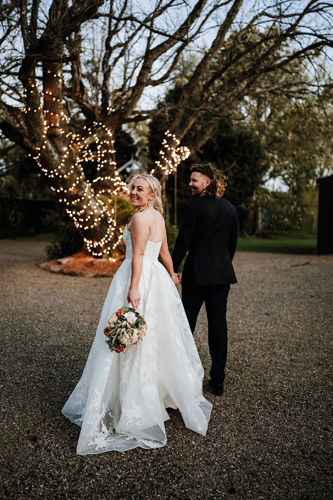 Bride in white gown holding bouquet, standing with groom beside a lit tree outdoors at dusk