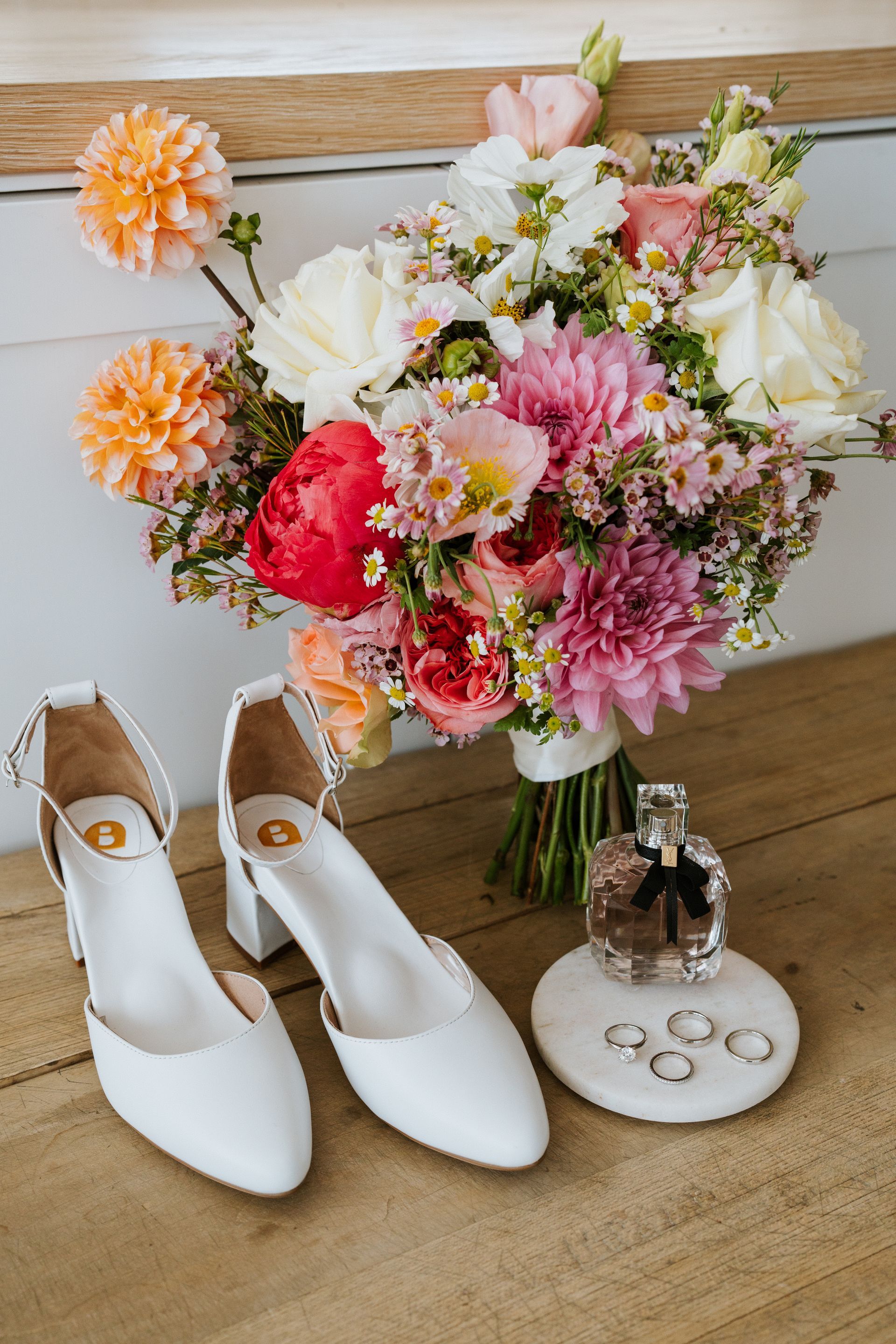 White bridal shoes beside a colorful bouquet and wedding rings on a wooden table