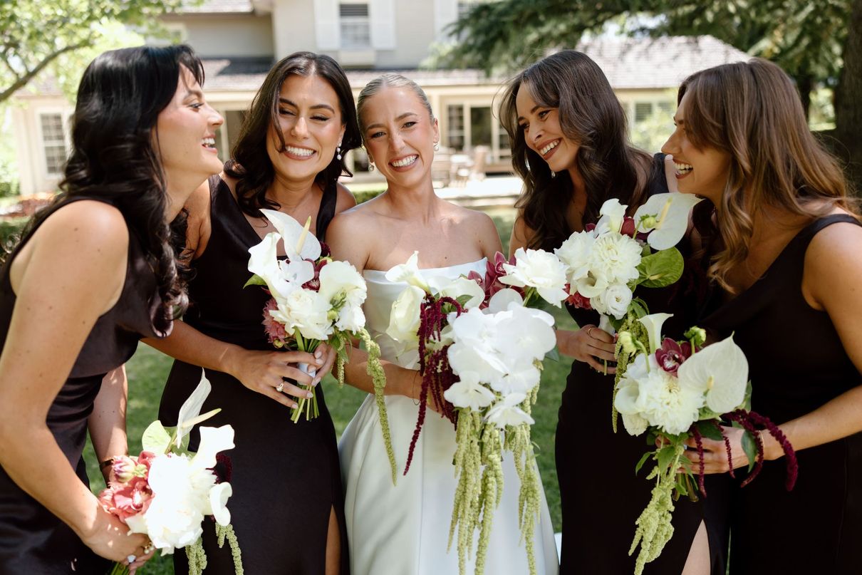 Five women in black dresses and a white dress smiling outdoors with bouquets, likely a bridal party