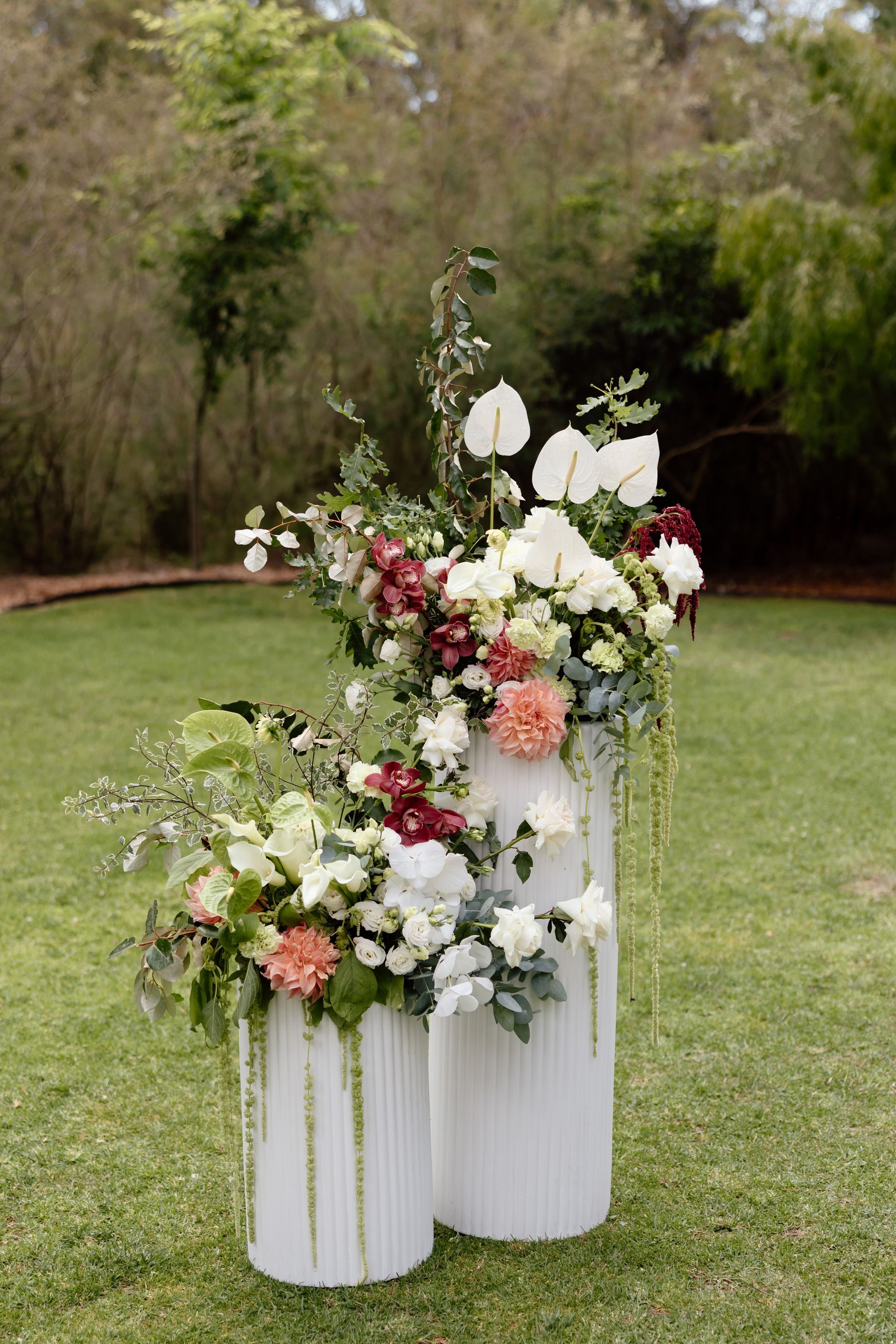 White floral arrangements with pink and green accents in tall vases on a grassy outdoor lawn